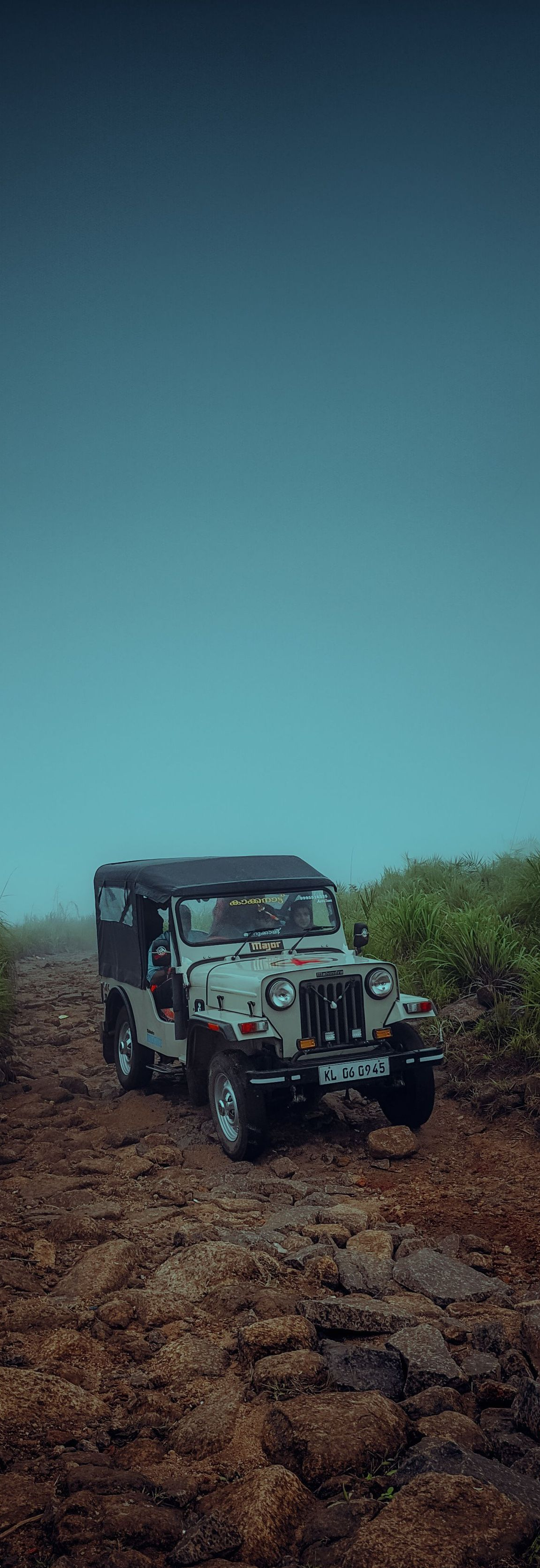 Off-road jeep driving on a rocky muddy trail beneath a cloudy blue sky.