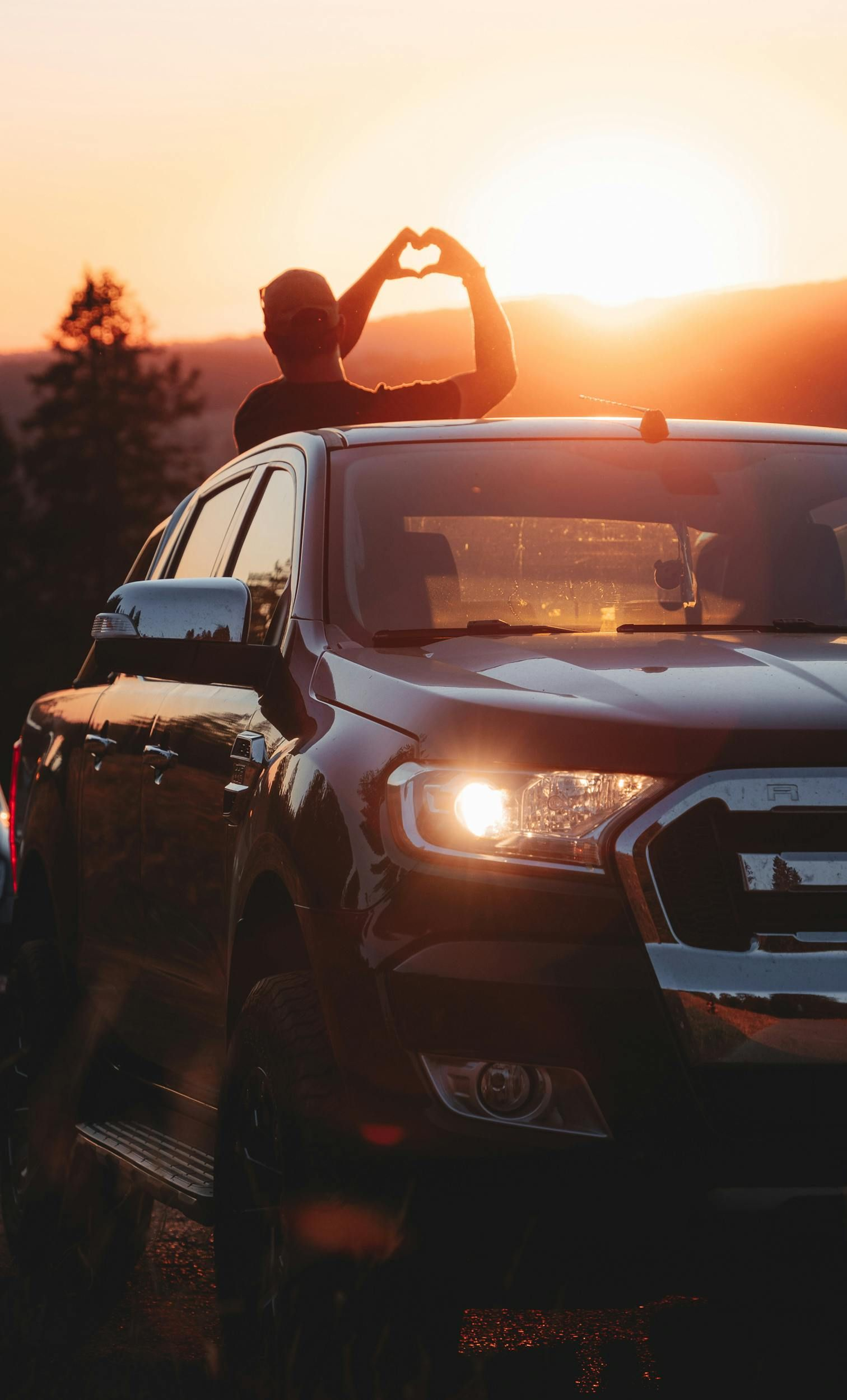Two people on a car rooftop at sunset, forming a heart shape with their hands.