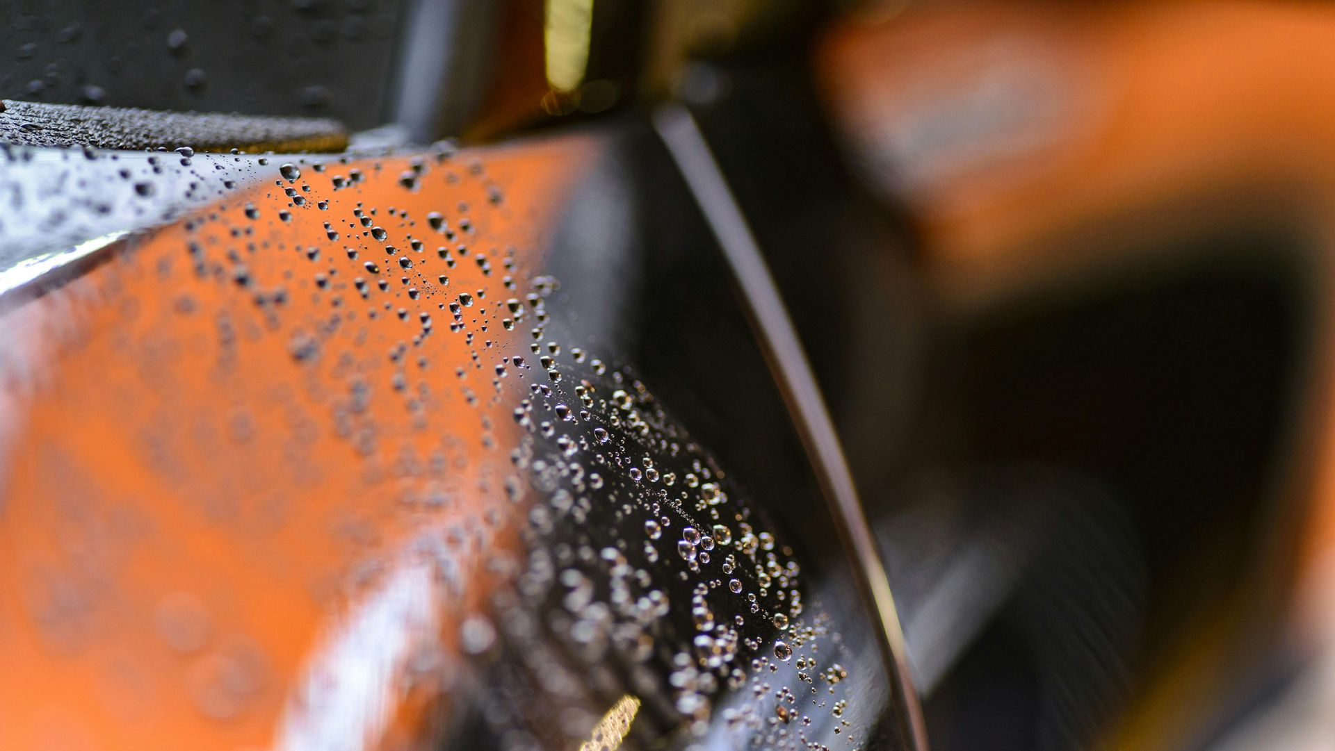 A close-up, angled view of a glossy orange vehicle surface covered in small raindrops.
