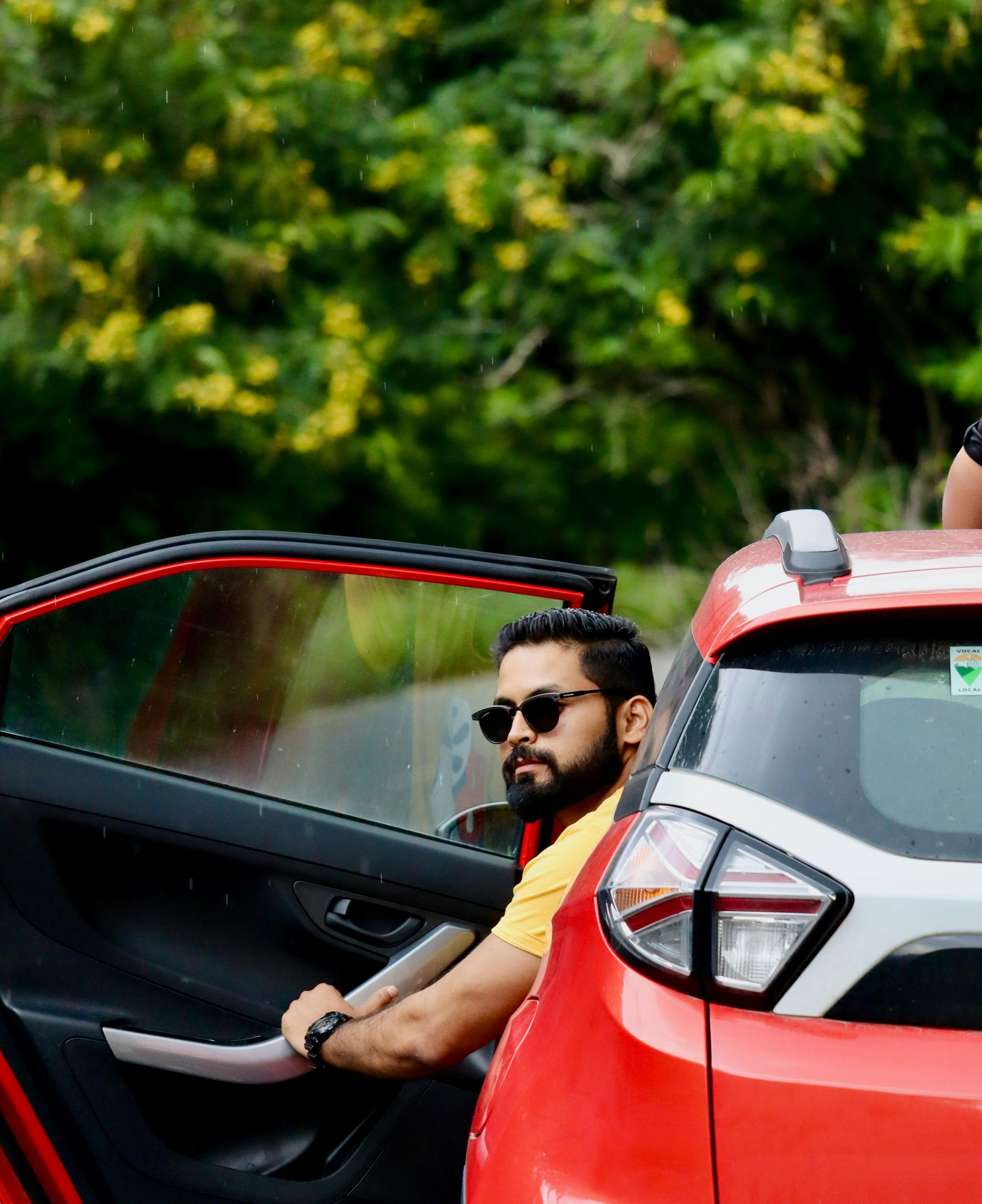 Man seated in a red car with the door open, wearing sunglasses, against a green leafy background