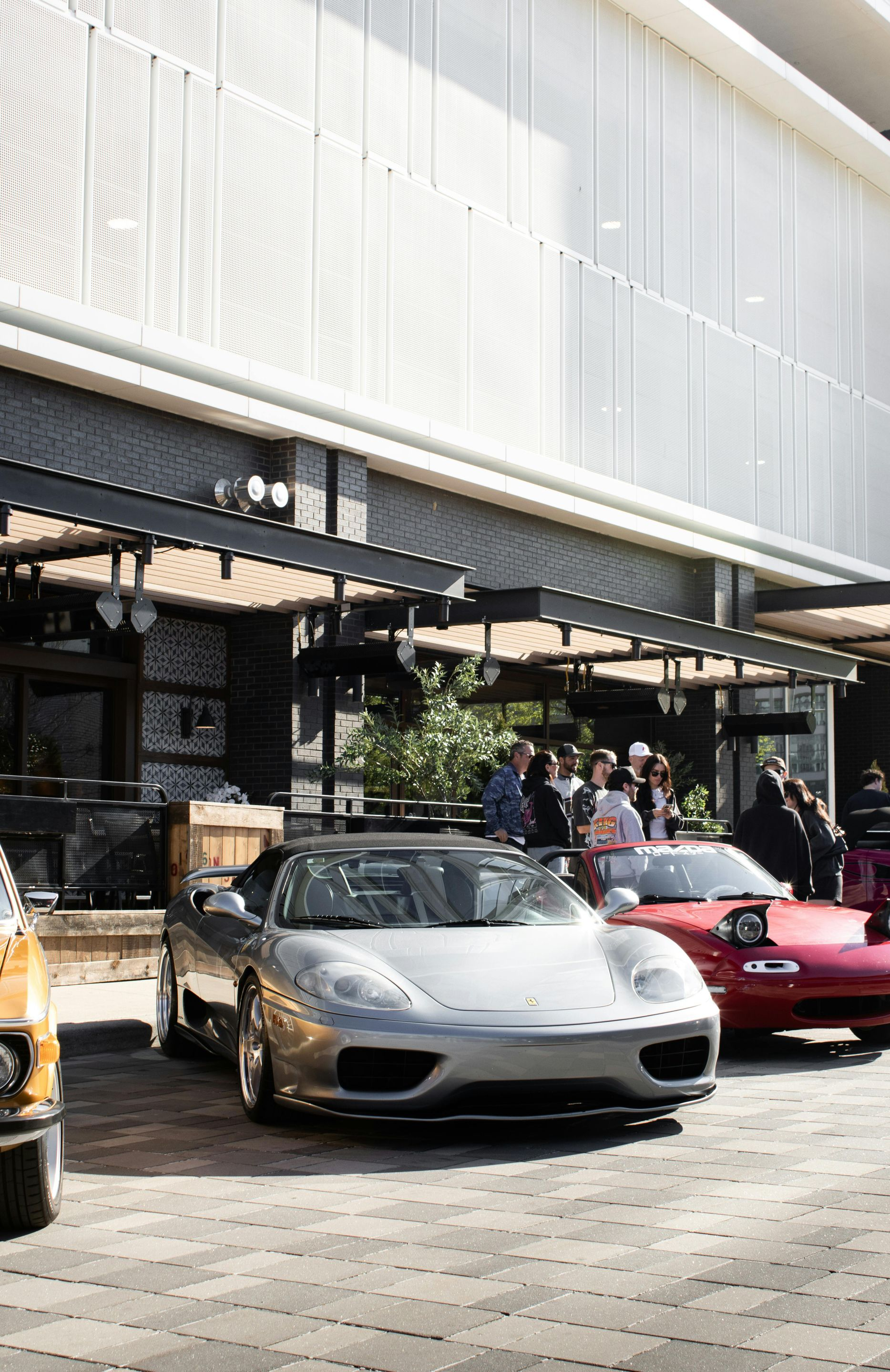 Vintage cars parked outside a modern building, with people gathered under an awning.