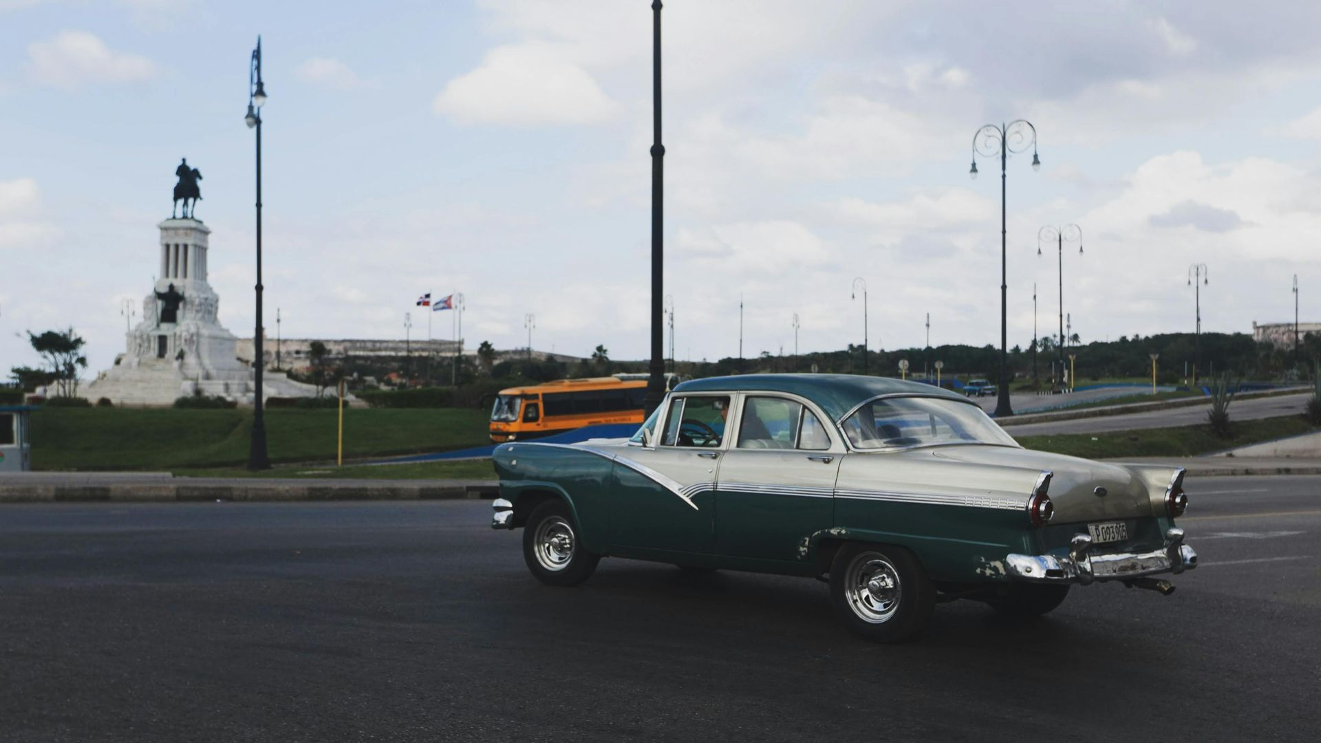 Classic green-and-white car driving on a roadway near a waterfront monument and lampposts