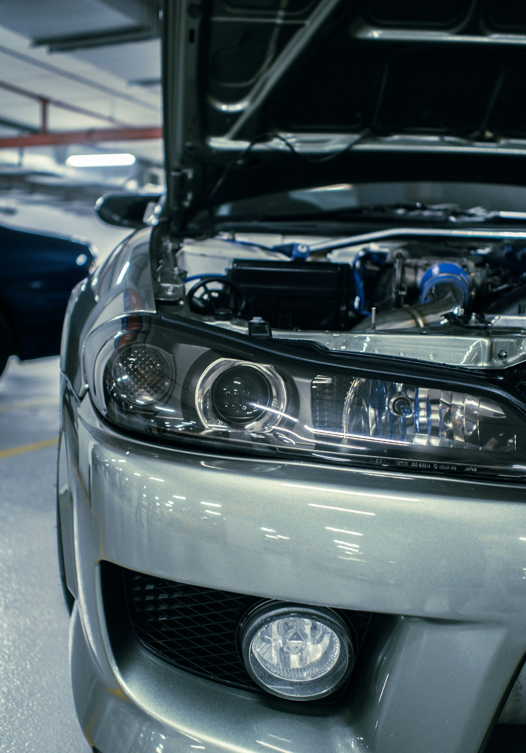 Silver car with open hood in a parking garage, showing the front headlight and fog light.