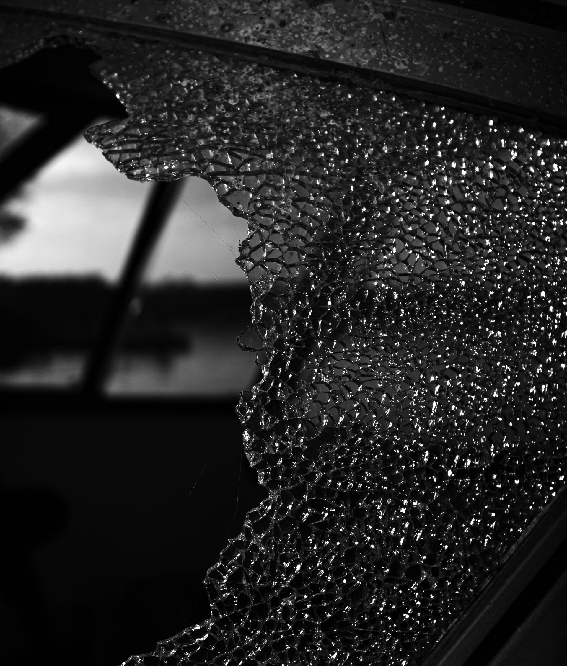 A close-up, black-and-white view of a shattered glass pane, looking out onto a blurry outdoor scene.