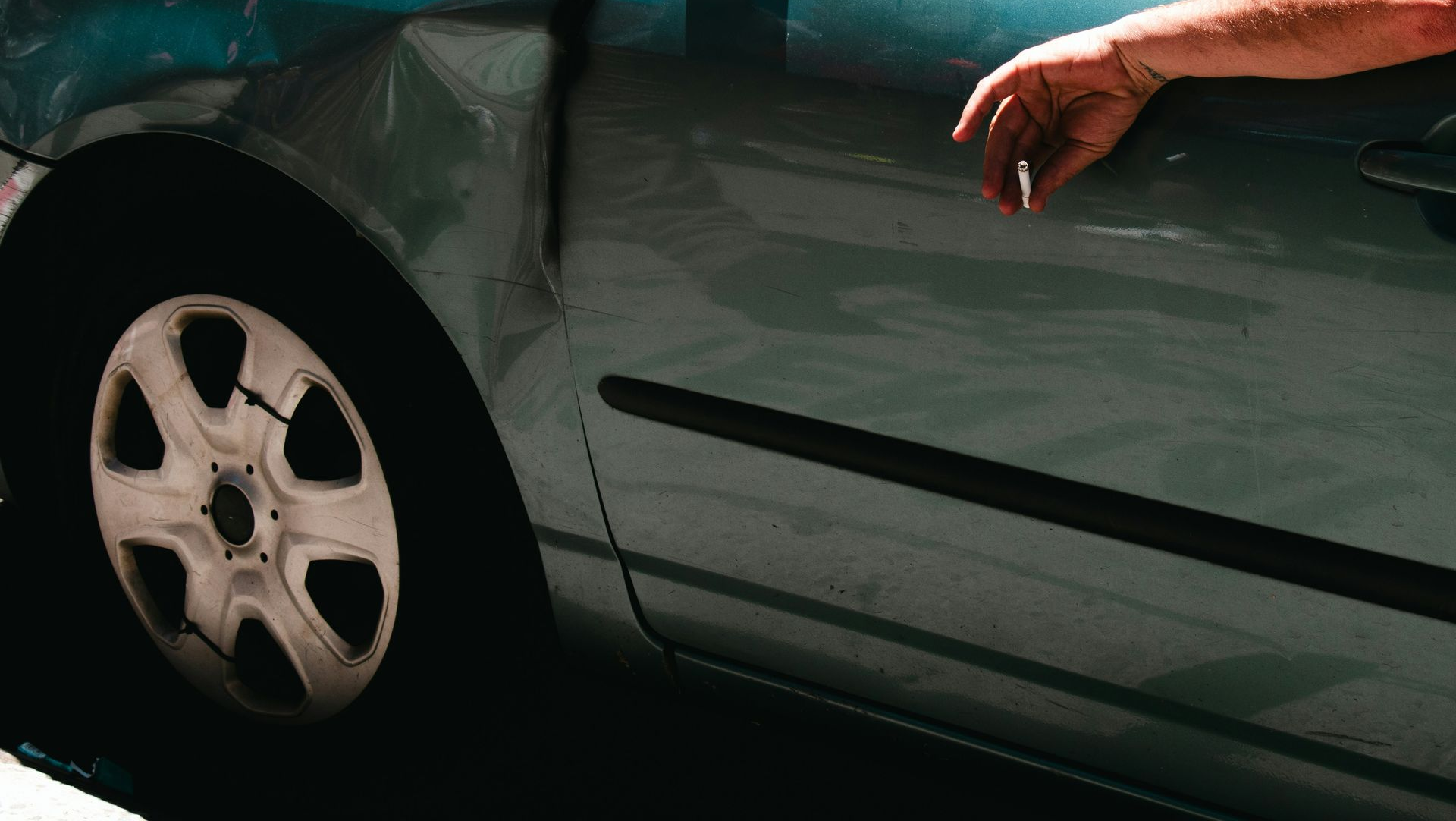 Damaged teal car with a dented front end and a person’s arm reaching from the window