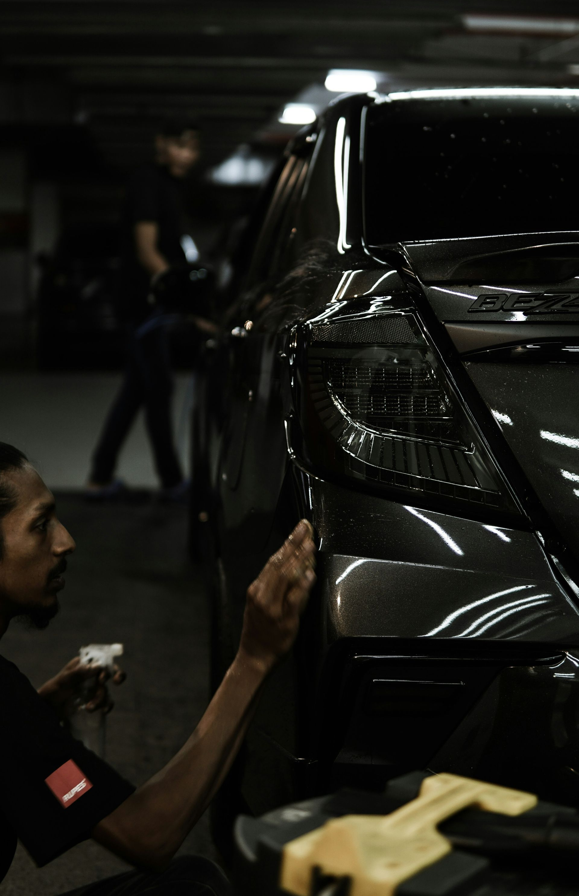 Detailing technician polishing a black car in a dim garage, with another person blurred in the background.