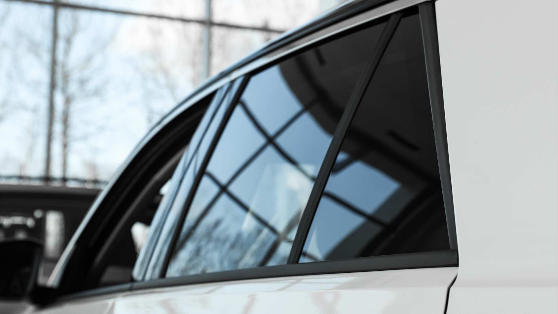 Close-up of a white car's side window, tinted dark, reflecting a building with glass windows.