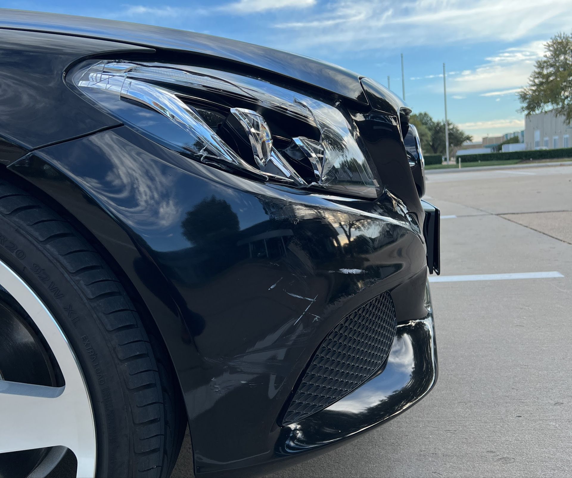 Close-up of a black car’s front bumper and headlight in a parking lot under a blue sky.