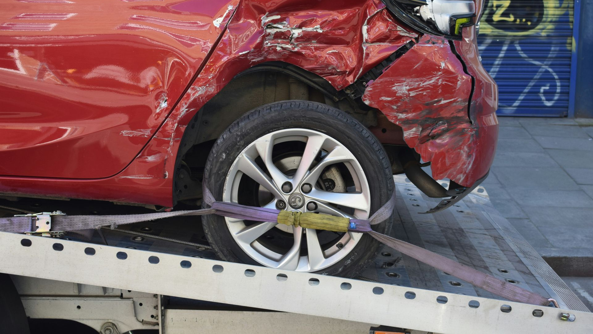 A damaged red car secured with tie-down straps on a flatbed tow truck.