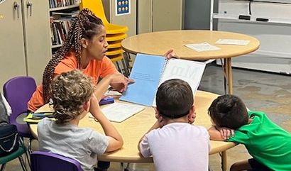 A person reads from a book to three children seated around a round table indoors.