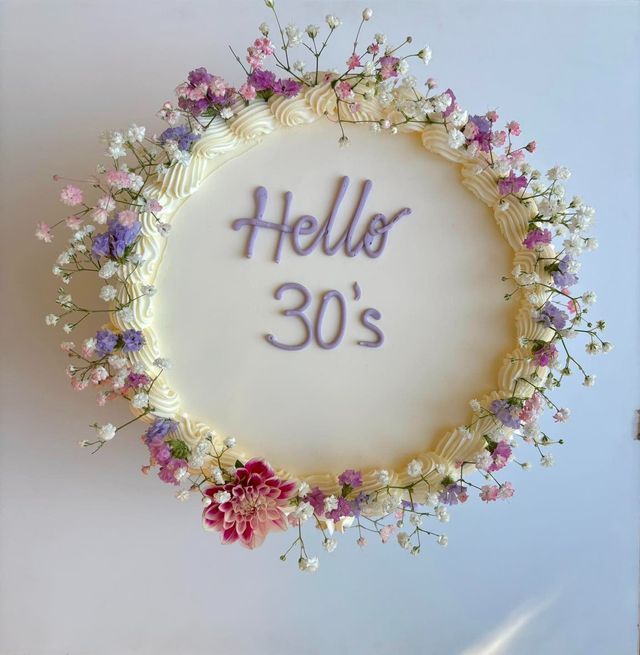 Torta decorata con fiori, scritta "Hello 30's" con glassa viola su una torta glassata bianca.