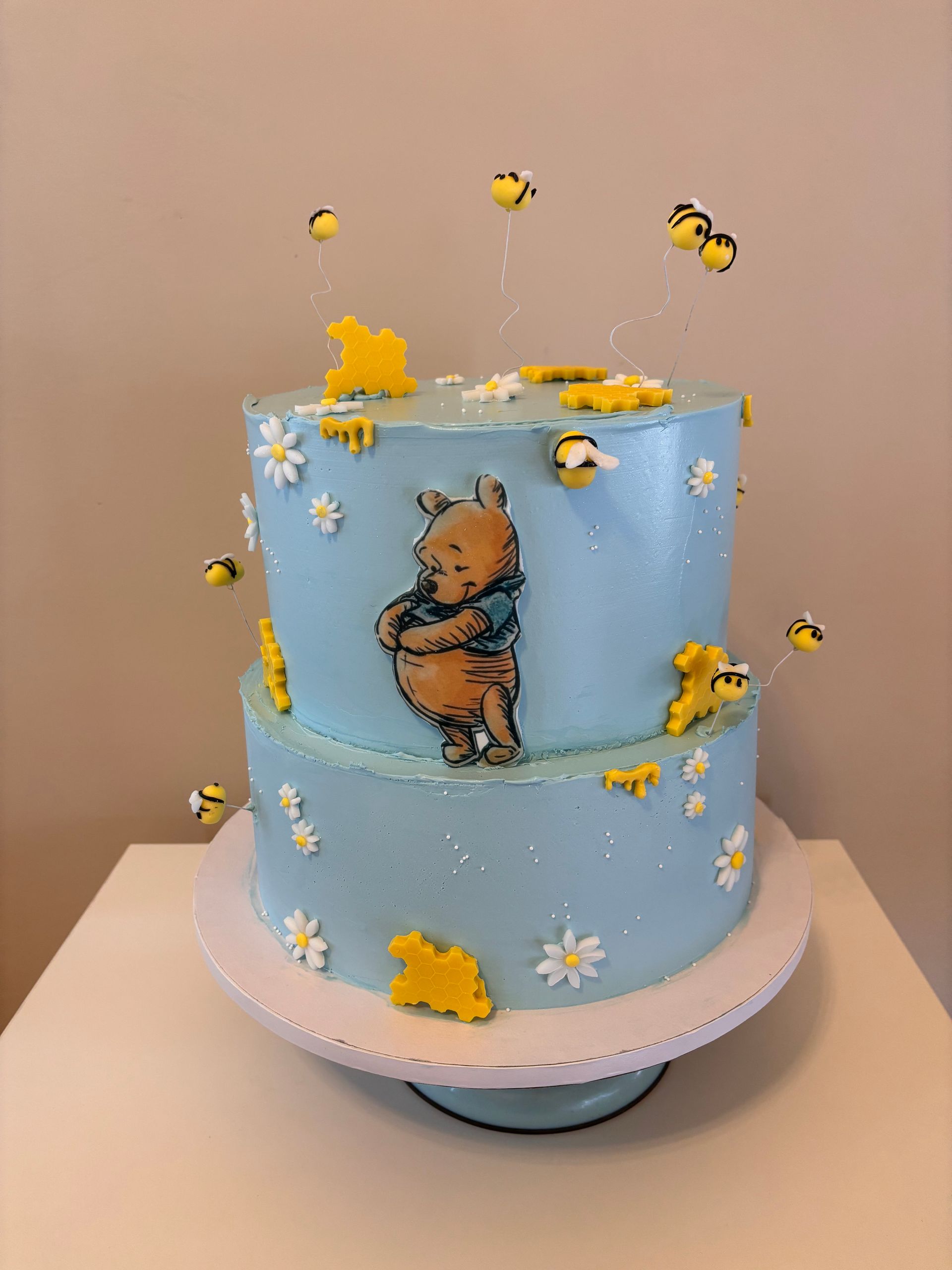 Torta a due piani con motivo Winnie the Pooh, decorata con api e fiori, su un'alzata bianca.