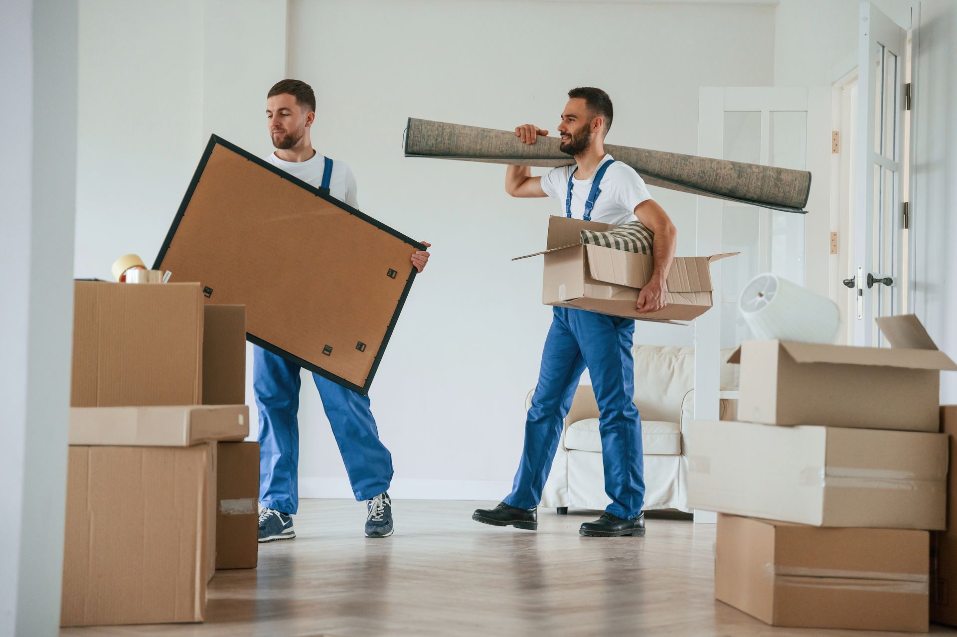Couple carrying a large cardboard box in a room with other boxes, hardwood floors, and white walls.