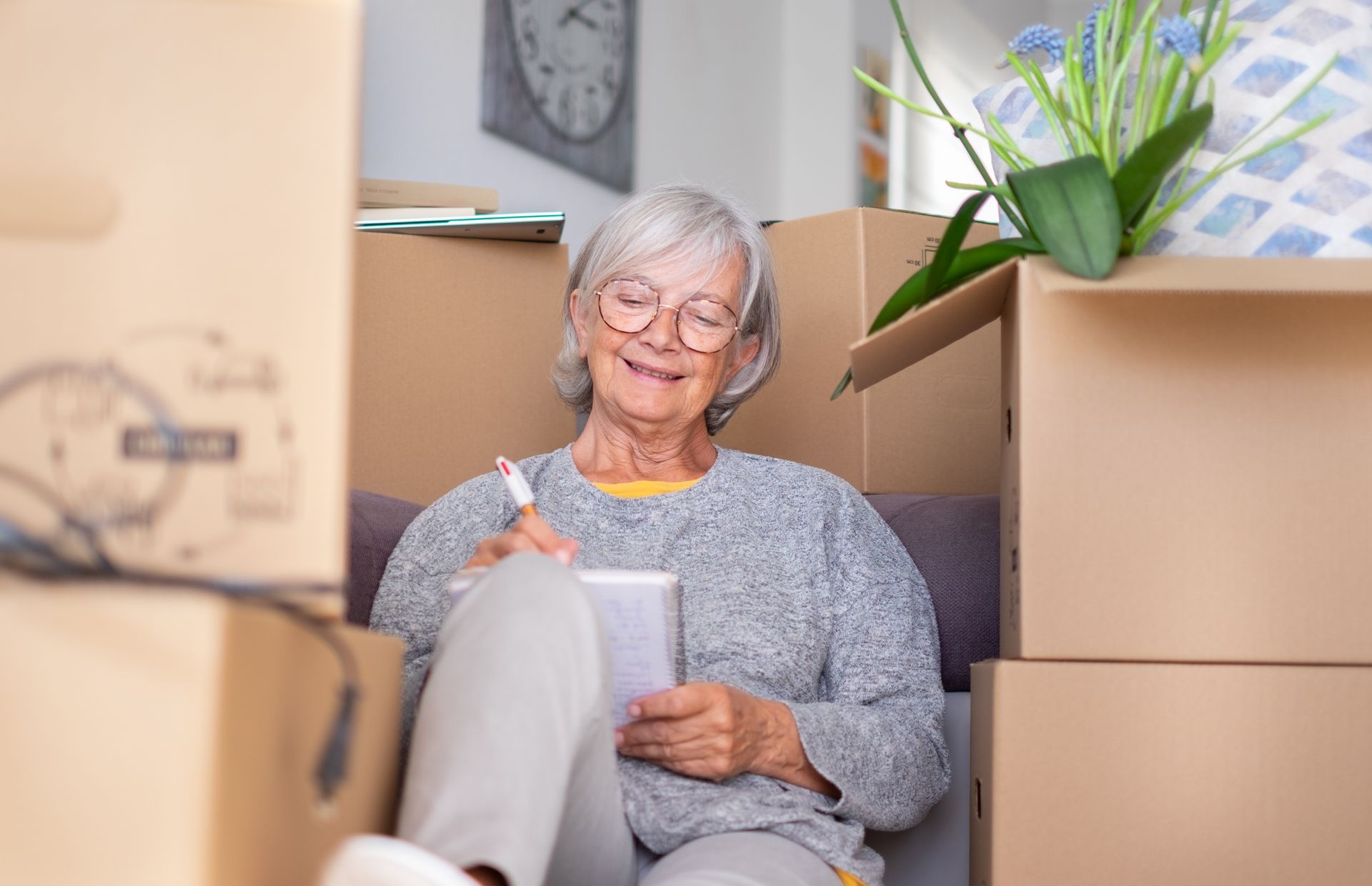 Elderly person sits among moving boxes, writing in a notebook, smiling.