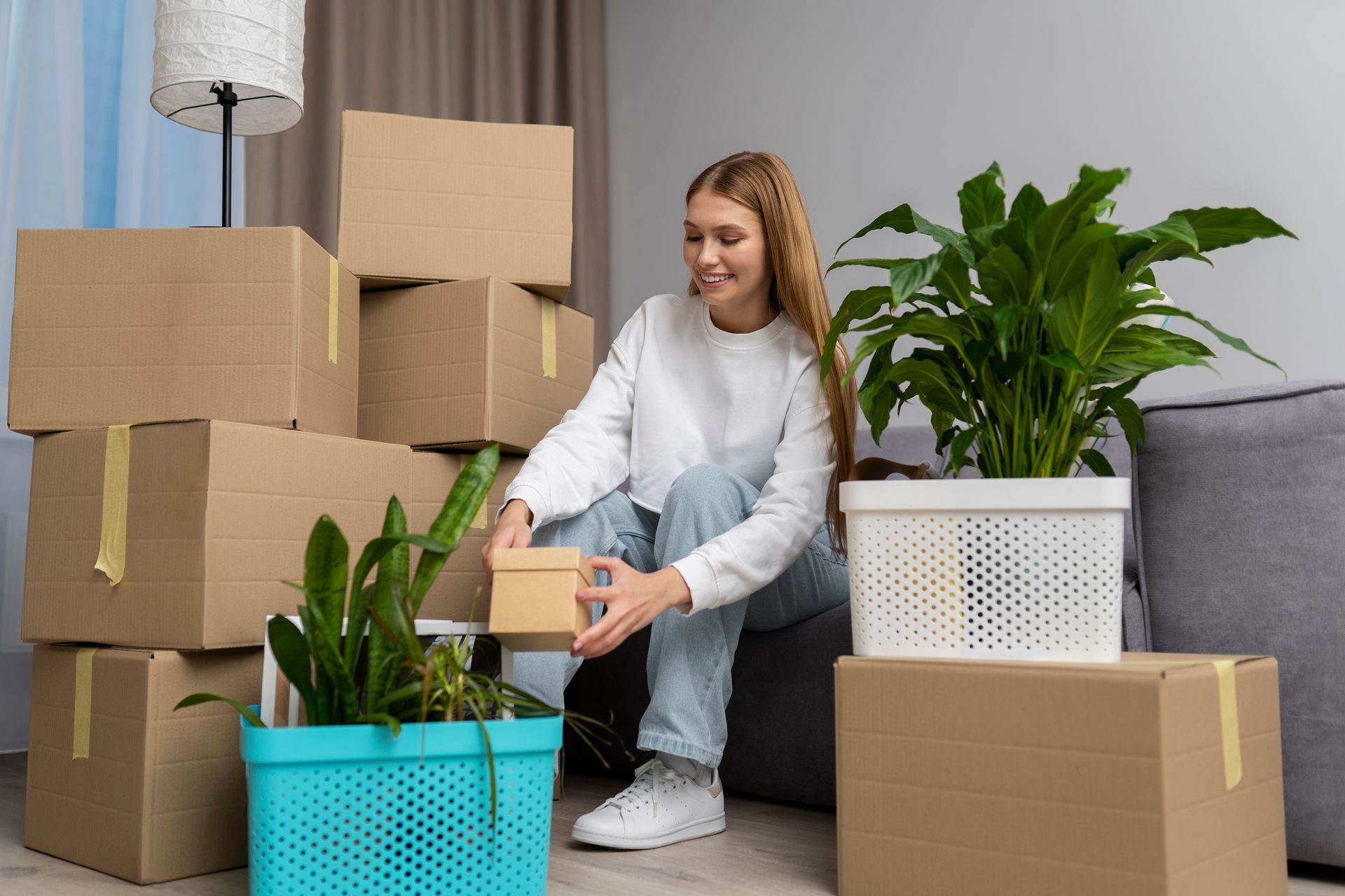 A woman is sitting on a couch surrounded by boxes and plants.