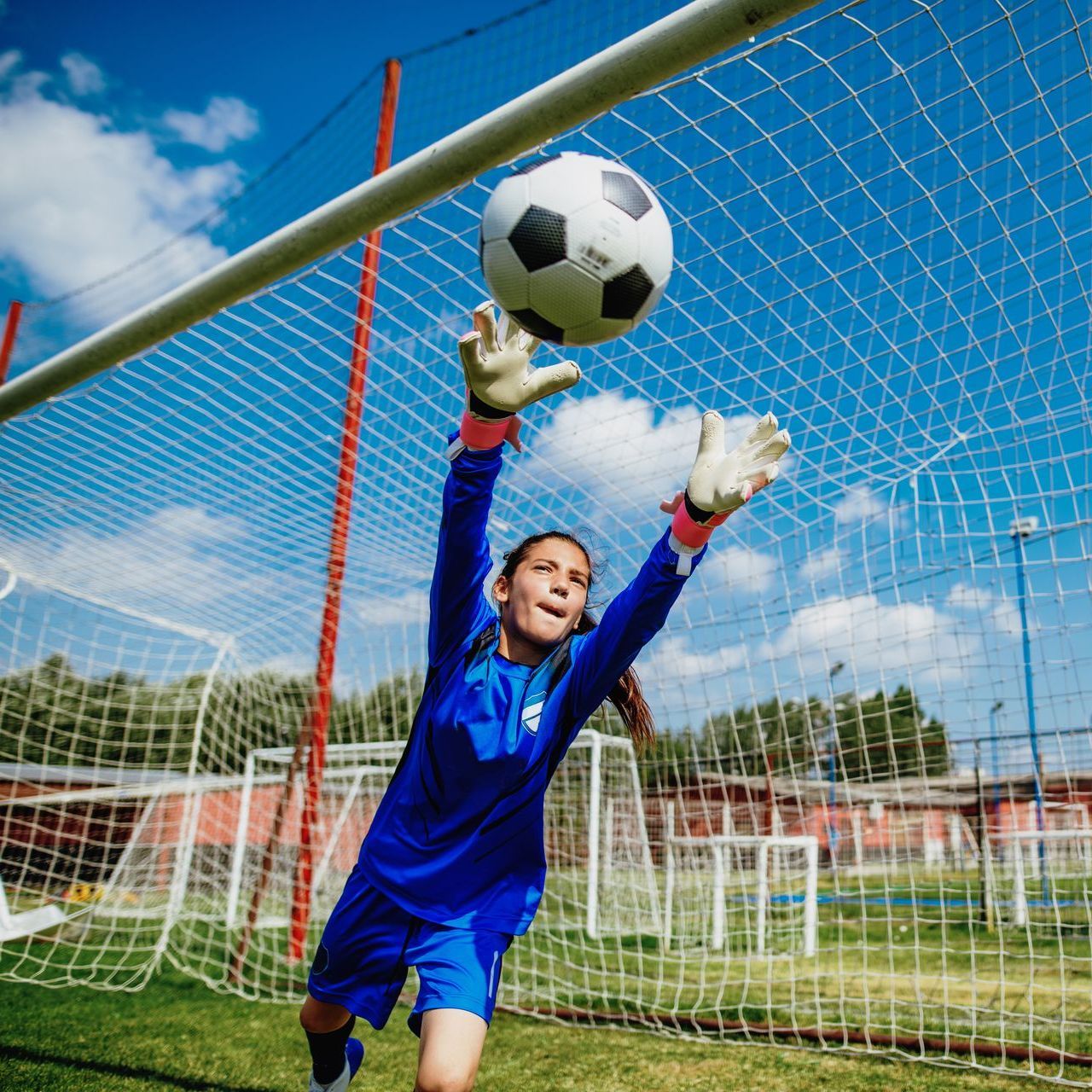 A young female soccer goalie is jumping to catch a soccer ball.
