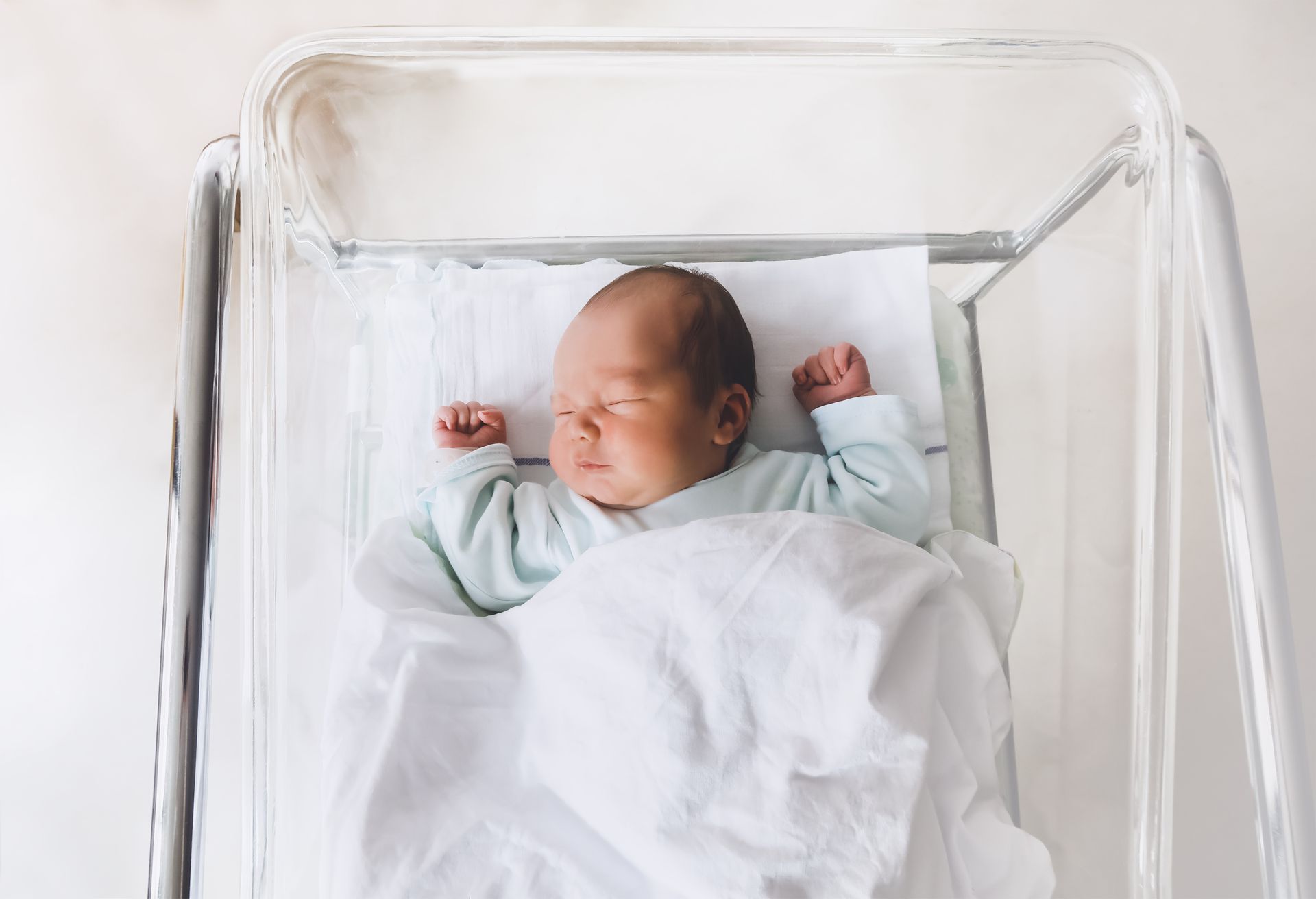 A newborn baby is sleeping in a clear crib.