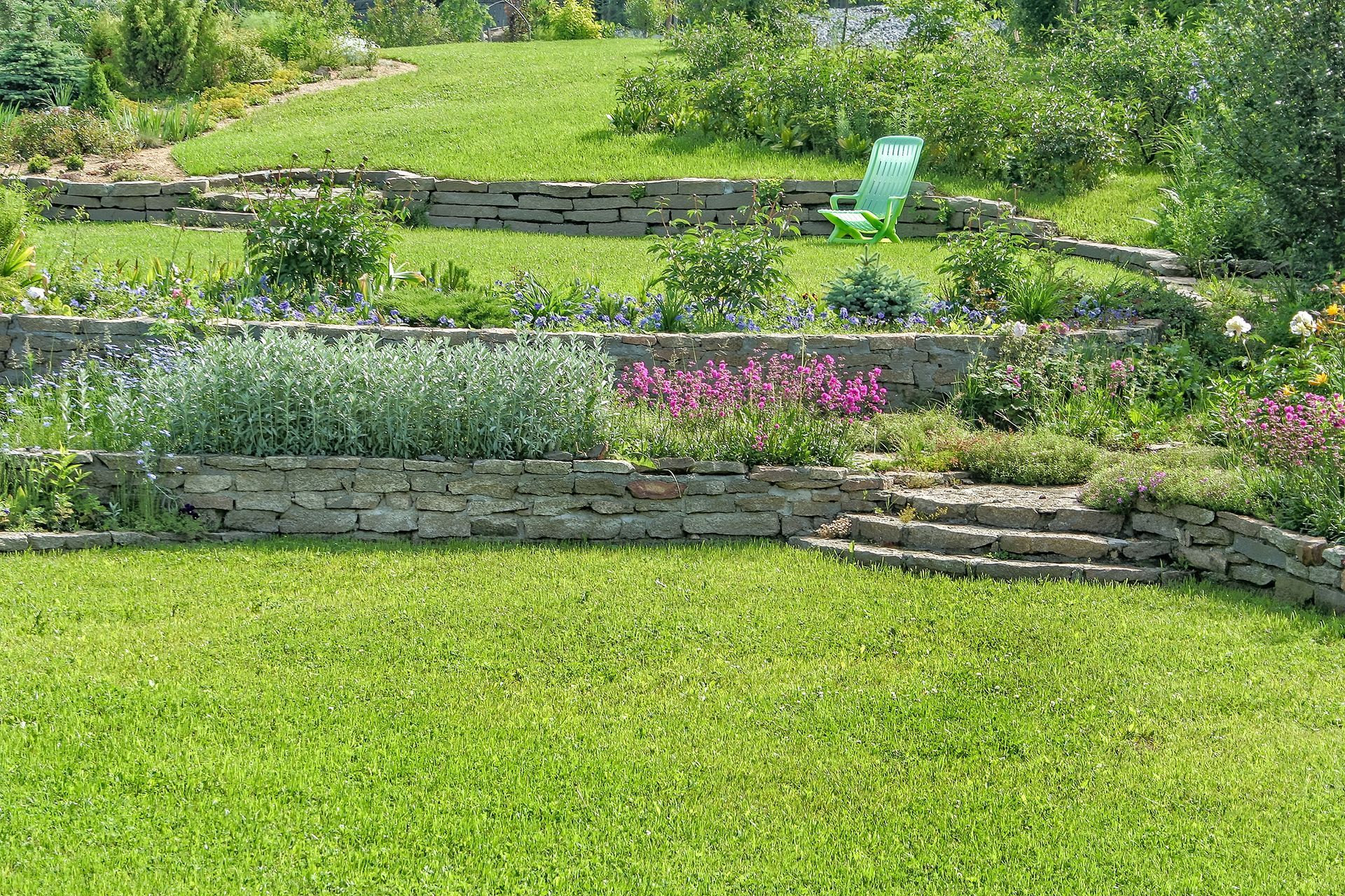 A tiered garden with stone retaining walls, vibrant flowers, green bushes, and a bright green chair on a grassy slope.