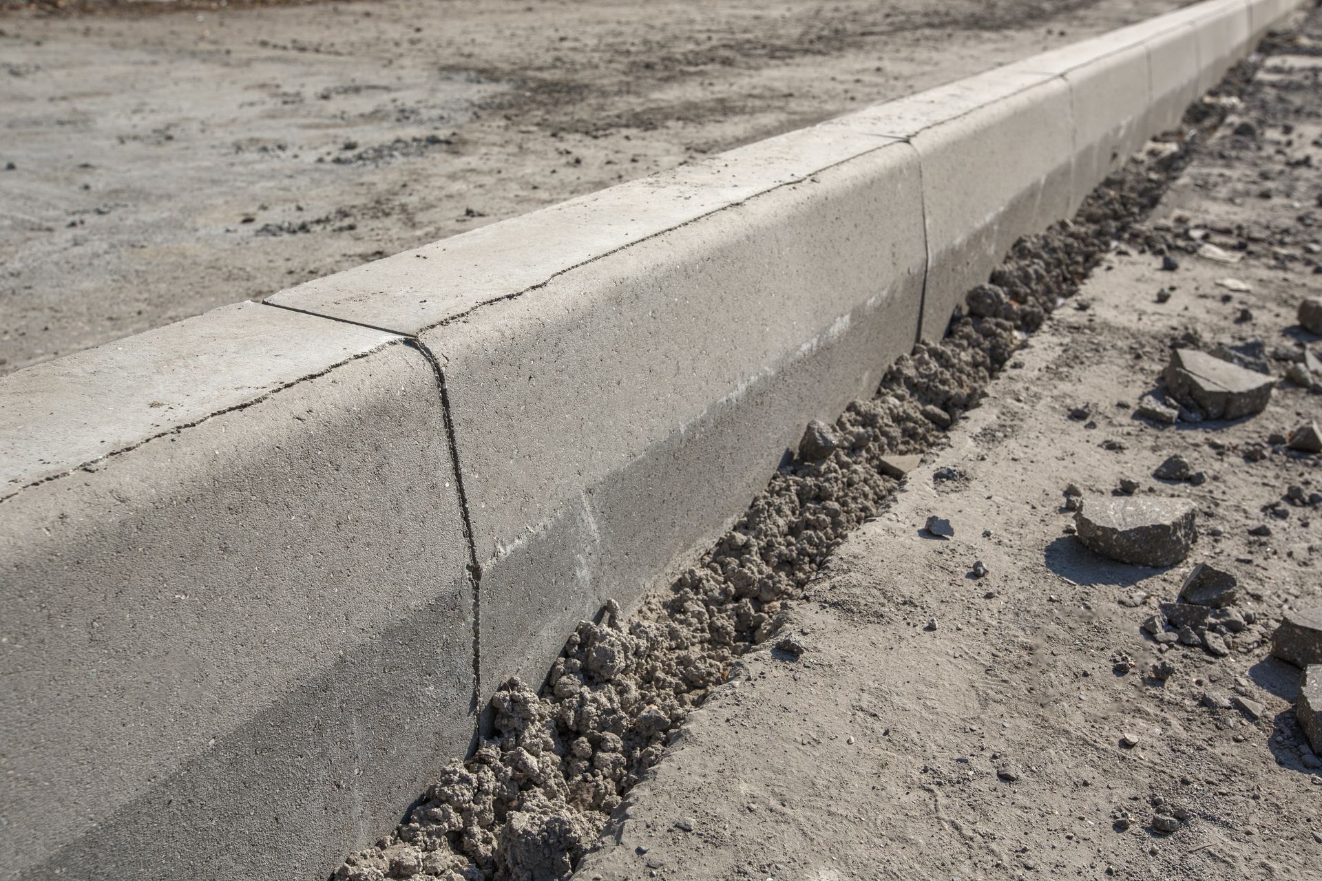 Gray concrete curb segments installed along a dusty, unpaved construction site ground. Gray concrete curb segments installed along a dusty, unpaved construction site ground.