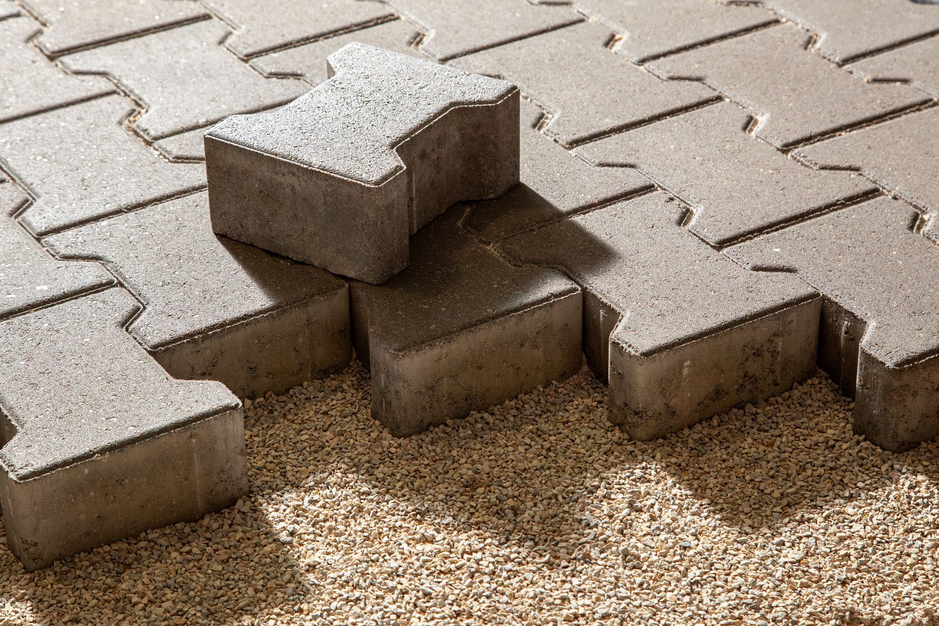 An I-shaped concrete paver sits on top of a partially laid gray interlocking pavement pattern over a gravel base. An I-shaped concrete paver sits on top of a partially laid gray interlocking pavement pattern over a gravel base.