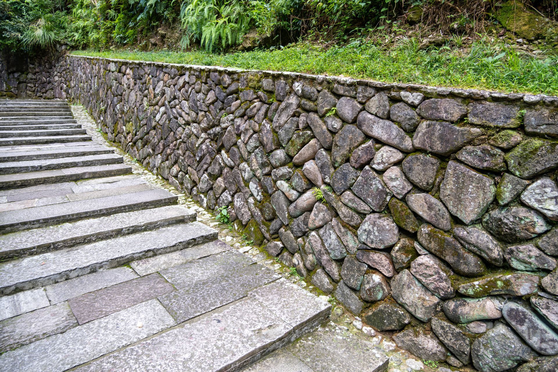 A stone stairway leading upward beside a rustic, mossy dry-stone retaining wall under a grassy embankment.