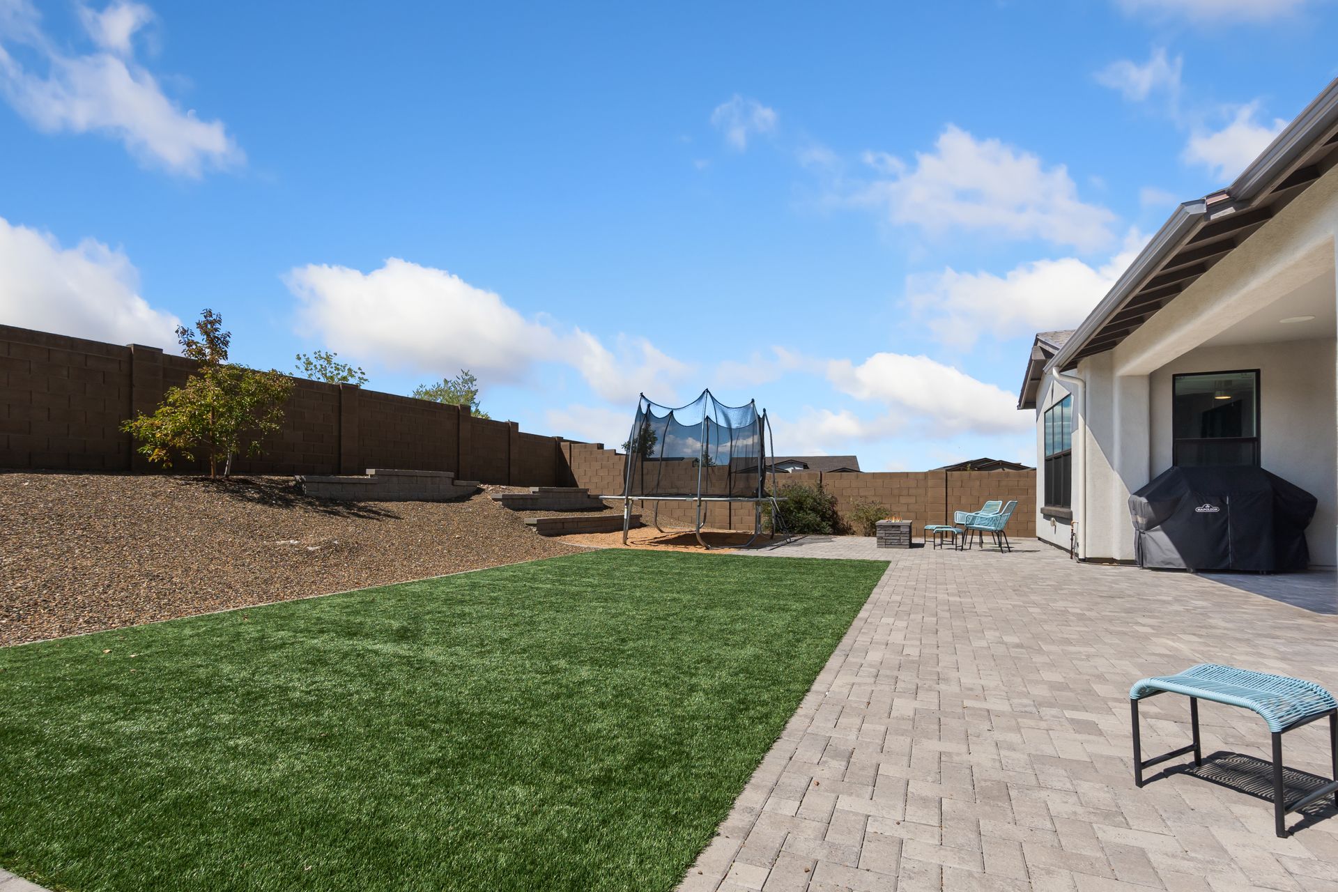 Backyard with green lawn, paver patio, trampoline, and covered house patio under a blue sky