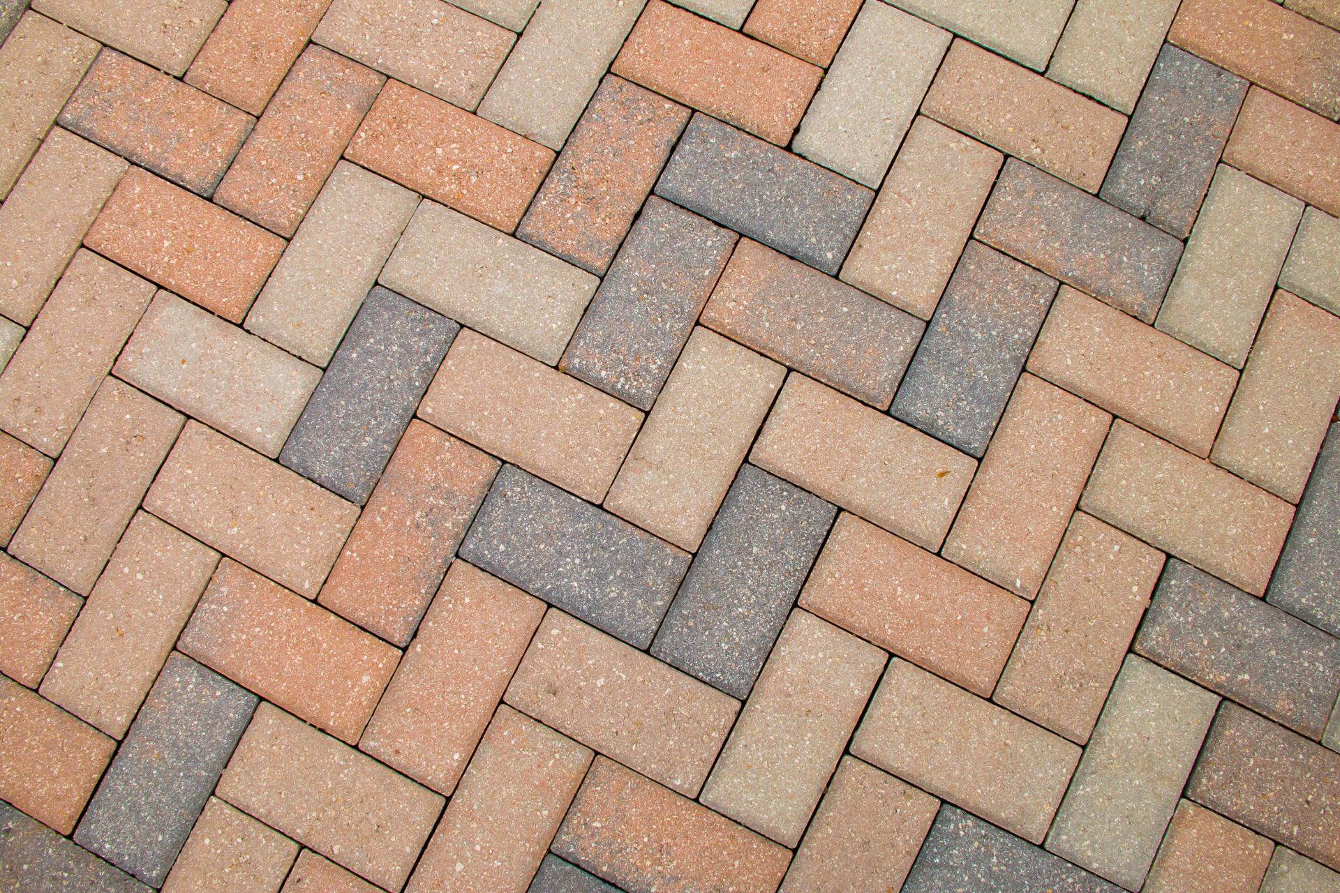 A herringbone pattern of rectangular paving stones in alternating shades of tan, brown, and grey. A herringbone pattern of rectangular paving stones in alternating shades of tan, brown, and grey.