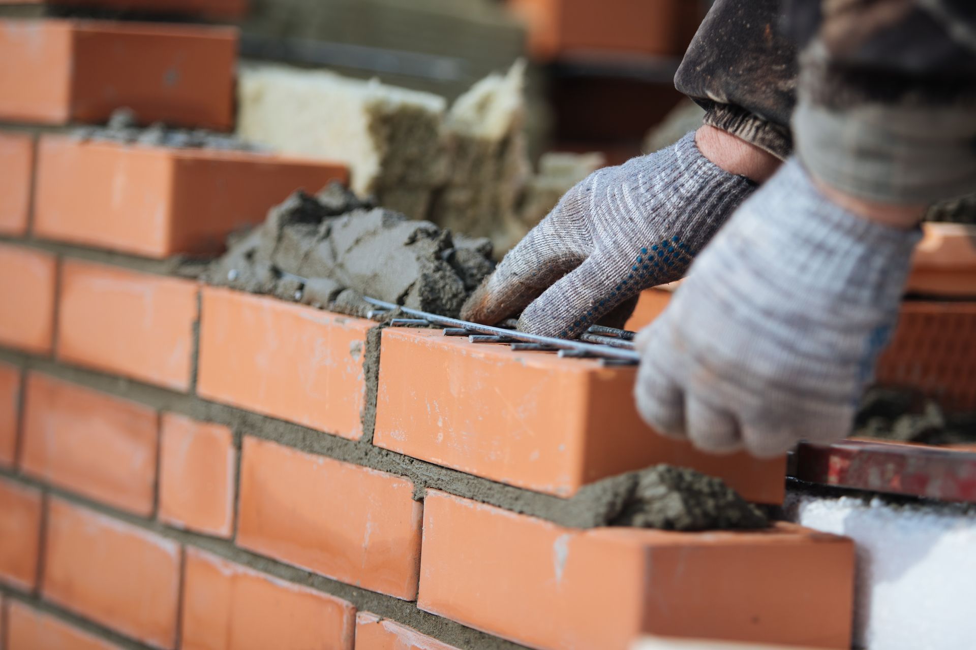 A worker in gray work gloves places metal reinforcement onto a layer of mortar between courses of red clay bricks. A worker in gray work gloves places metal reinforcement onto a layer of mortar between courses of red clay bricks.