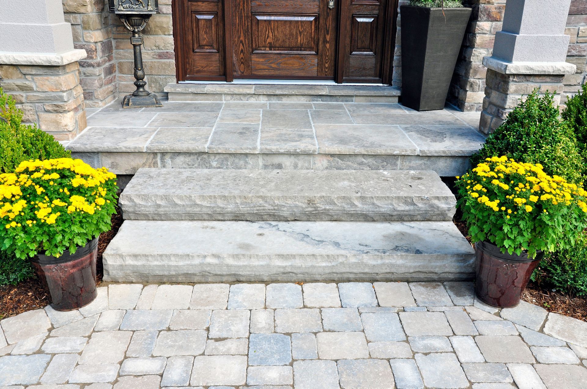 Stone steps leading to a front door, flanked by two potted yellow flower arrangements on a paved patio. Stone steps leading to a front door, flanked by two potted yellow flower arrangements on a paved patio.