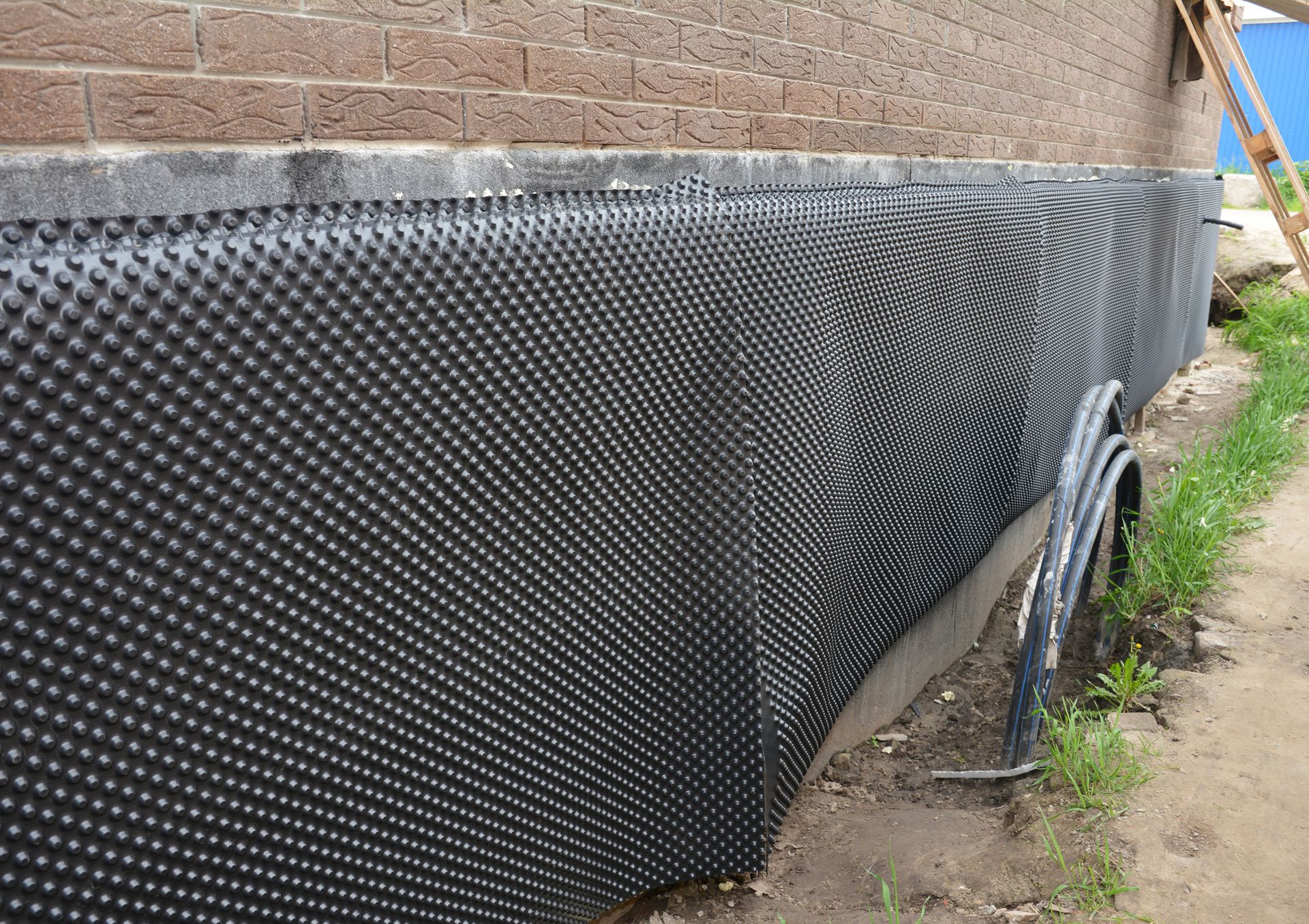 Black dimpled drainage membrane installed along a brick building foundation at ground level. Black dimpled drainage membrane installed along a brick building foundation at ground level.