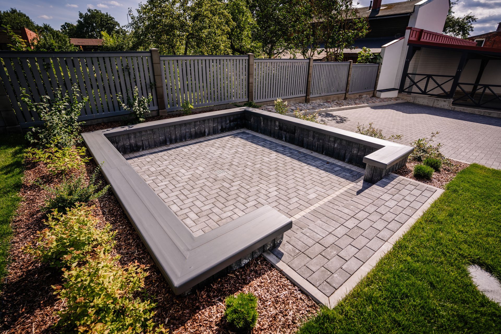 A sunken stone patio area with a built-in seating wall, surrounded by landscaping, gravel, and a fence in a backyard.