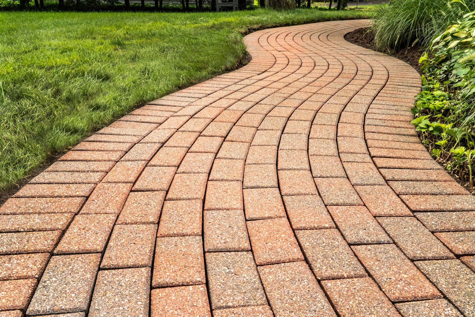 A winding path made of tan and reddish-brown rectangular paving stones, bordered by green grass and garden plants. A winding path made of tan and reddish-brown rectangular paving stones, bordered by green grass and garden plants.