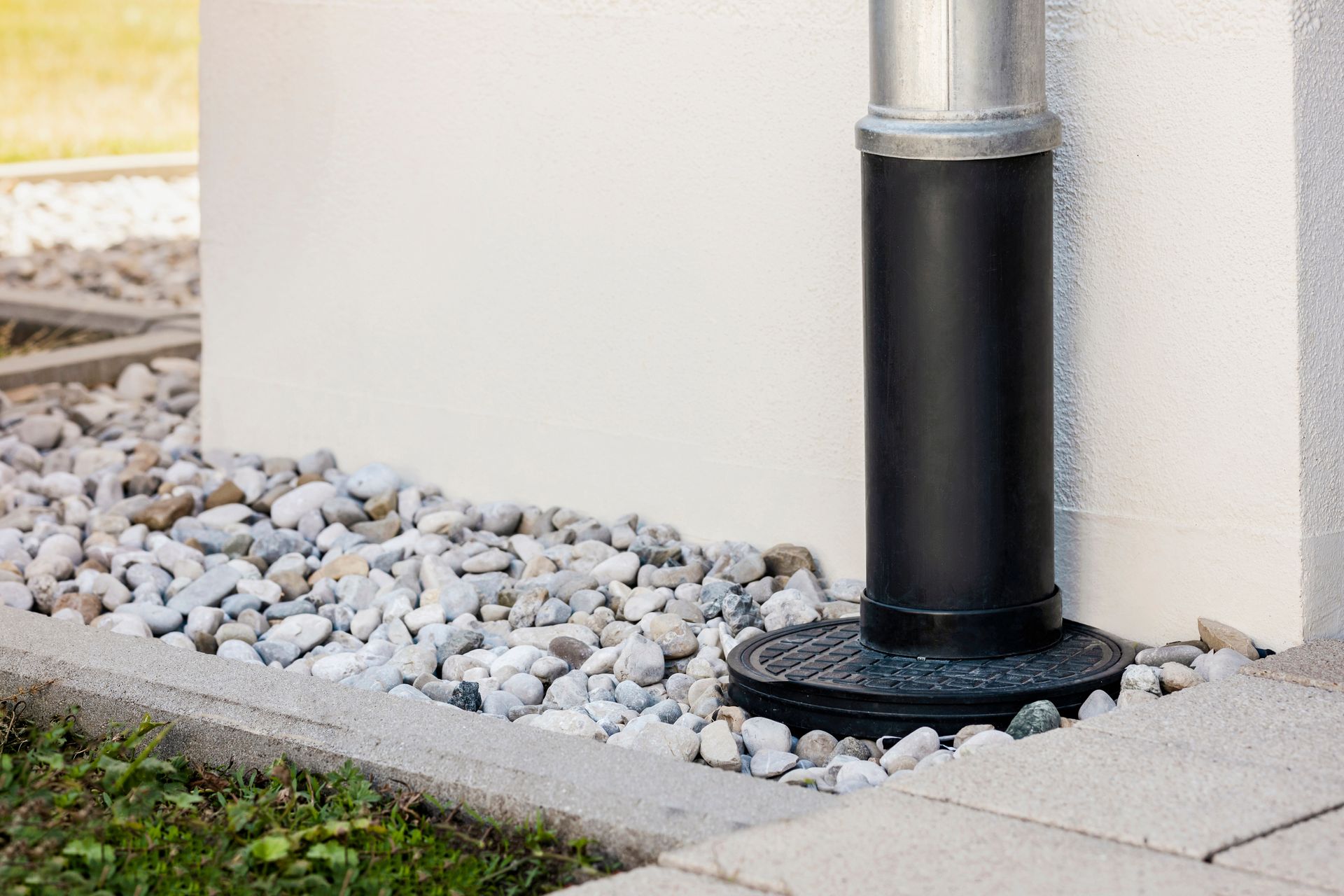 A metal and black pipe downspout connected to a ground-level drain, set against a light wall with a gravel border. A metal and black pipe downspout connected to a ground-level drain, set against a light wall with a gravel border.