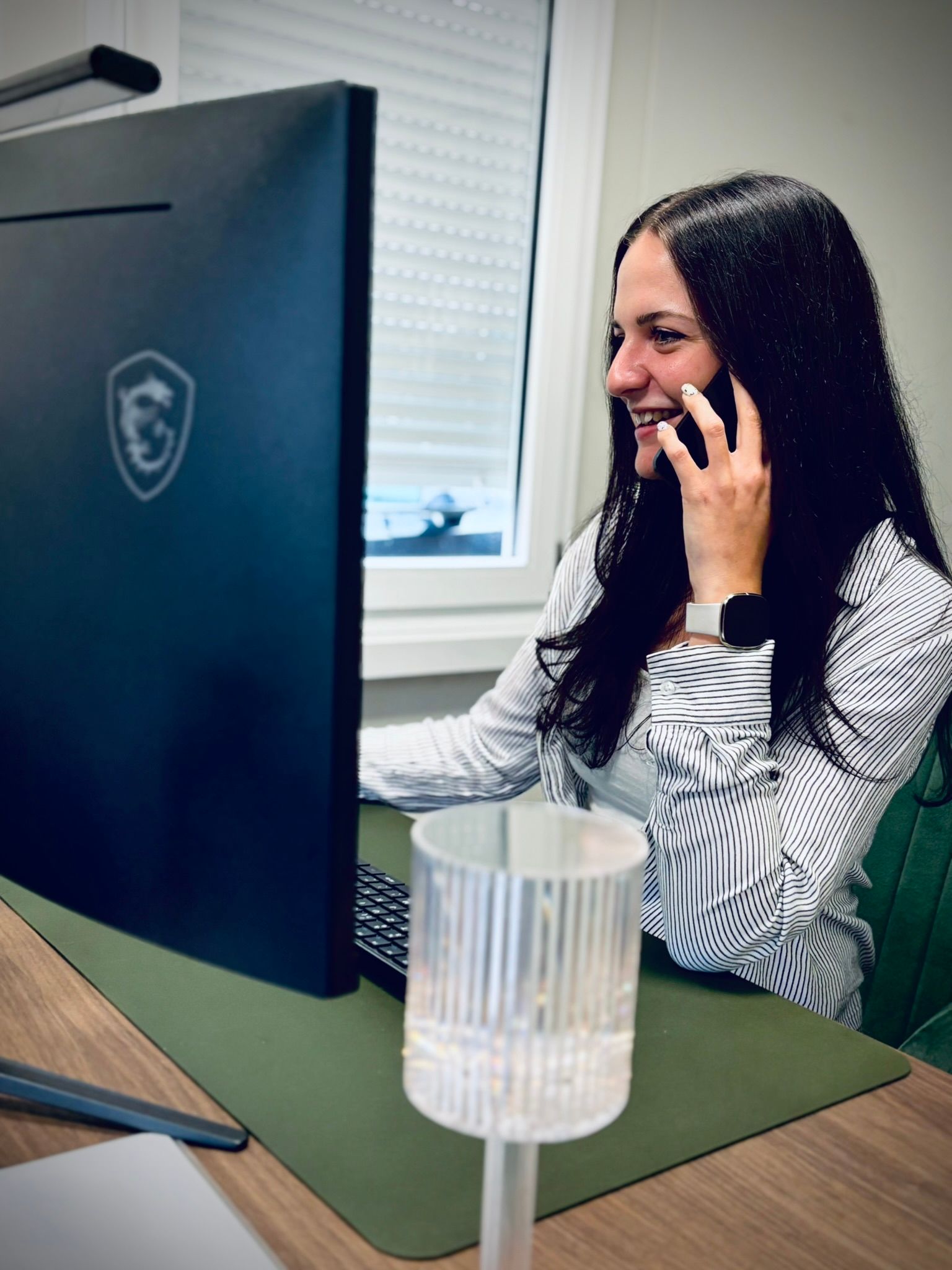 Eine Frau sitzt vor einem Computer und telefoniert mit einem Mobiltelefon.