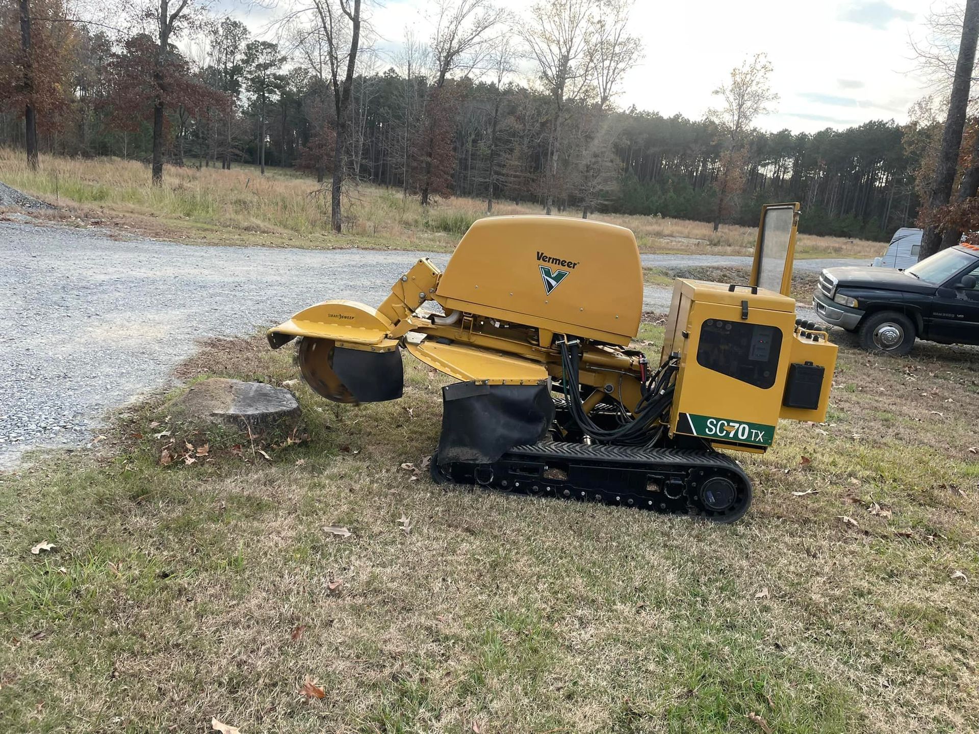 Yellow compact excavator on grass near a gravel road with trees in the background