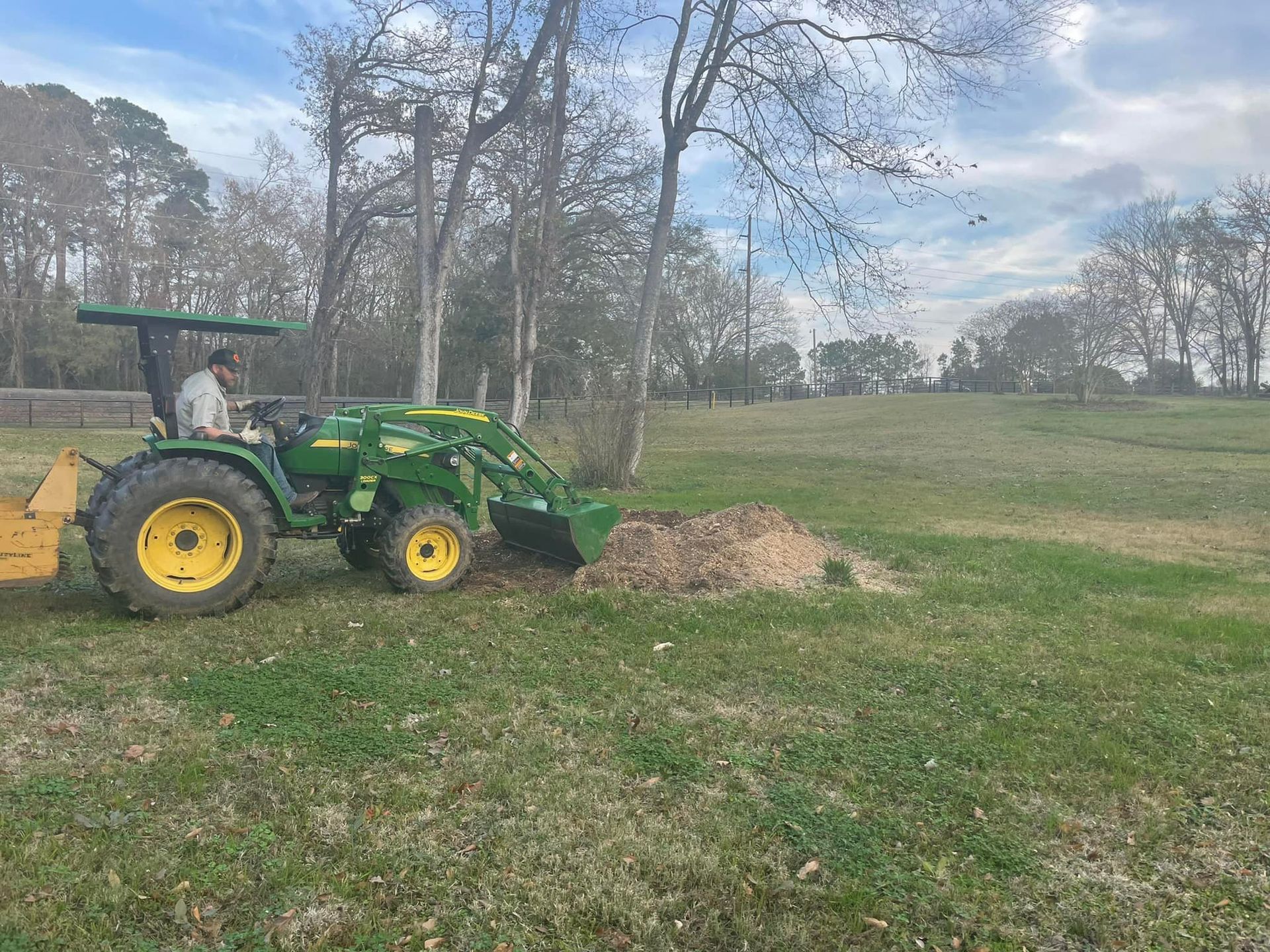 Green tractor with front loader pushing dirt in a grassy field near trees under a cloudy sky