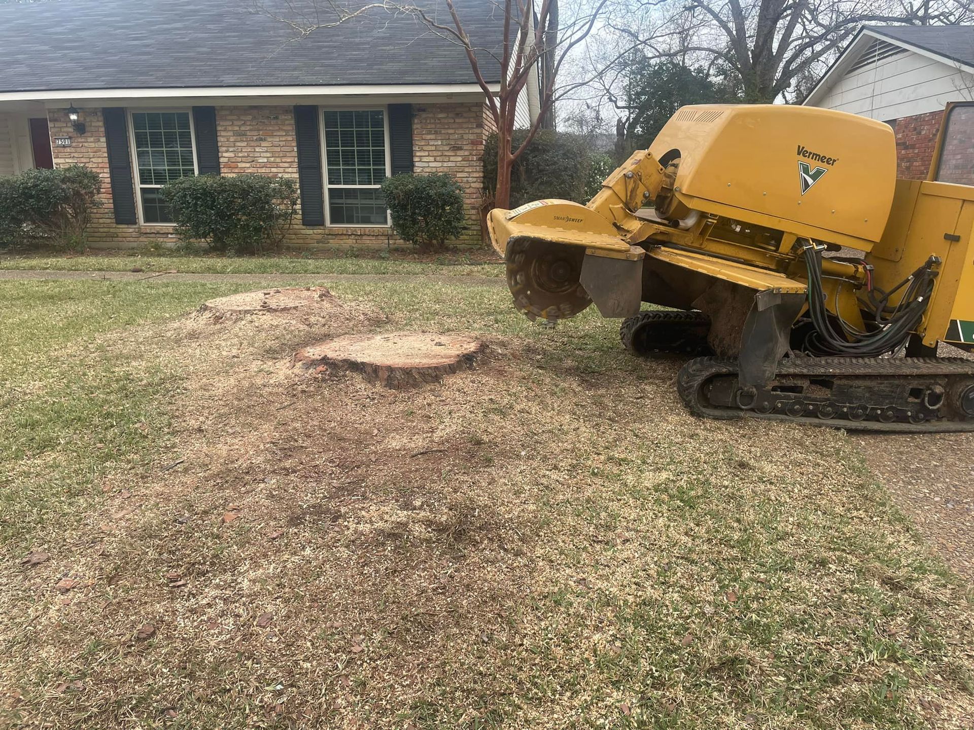 Yellow skid-steer near a brick house, moving soil in a yard with a bare patch of ground