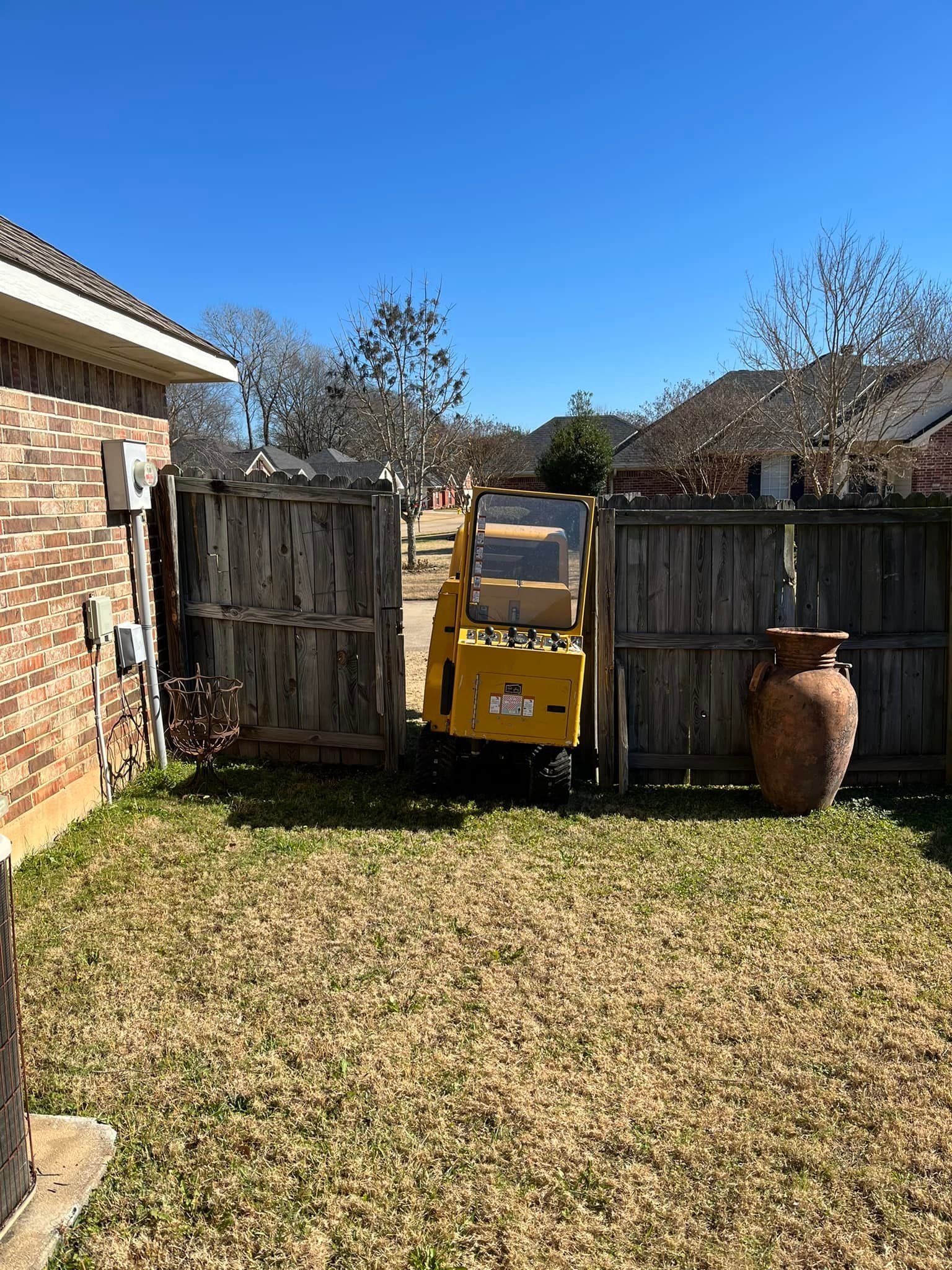 Yellow toy dump truck in a sunny backyard between wooden fences, with a brick house on the left and a large pot on the right