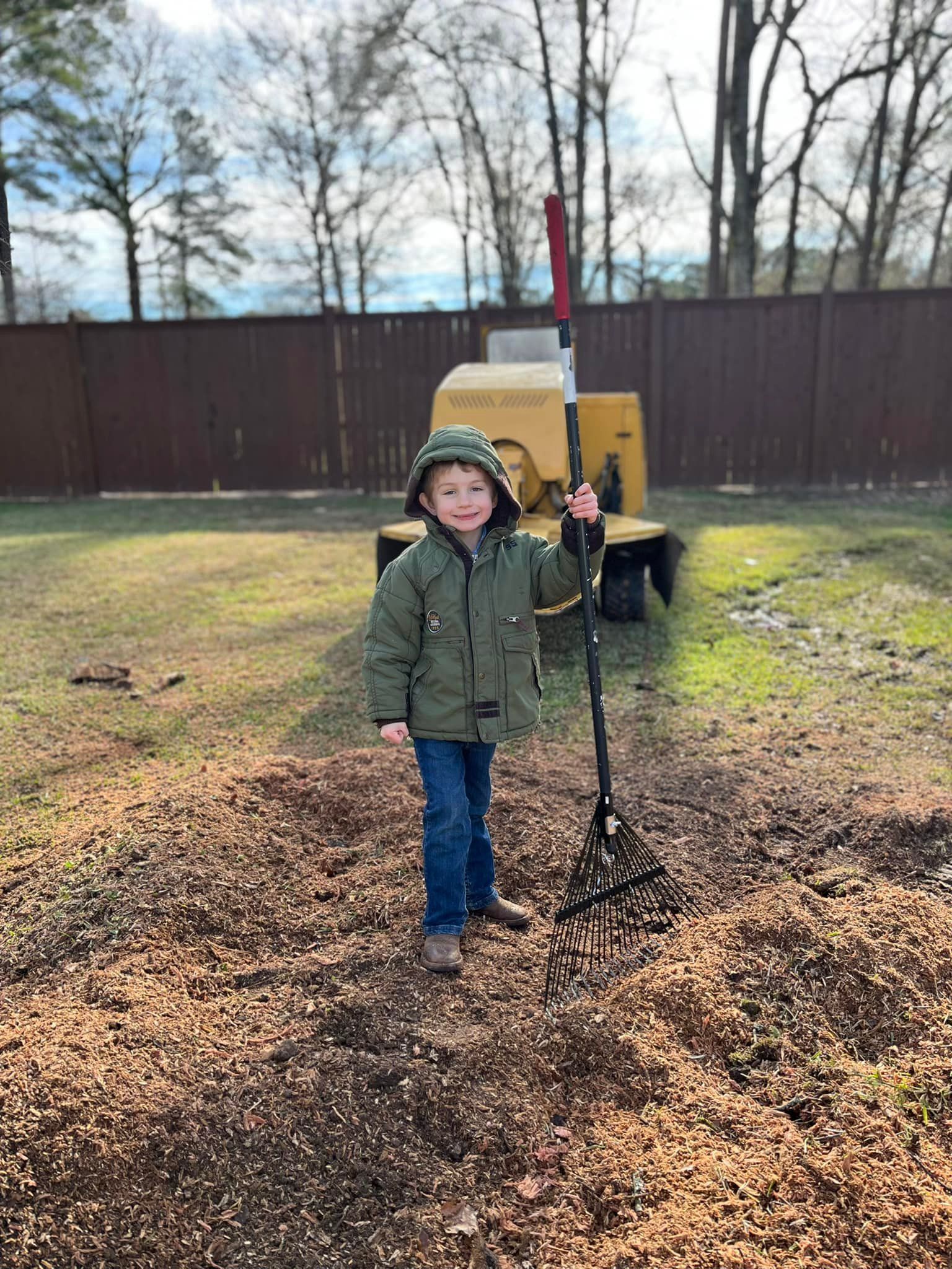 Child in green jacket stands by a tripod in a yard with trees, fence, and a yellow machine in the background.