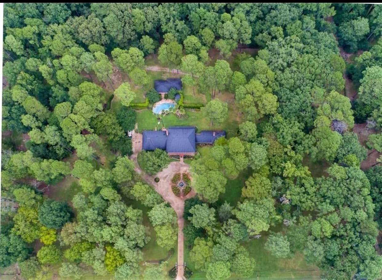 Aerial view of a house with a blue roof surrounded by dense green forest and a dirt path leading to it