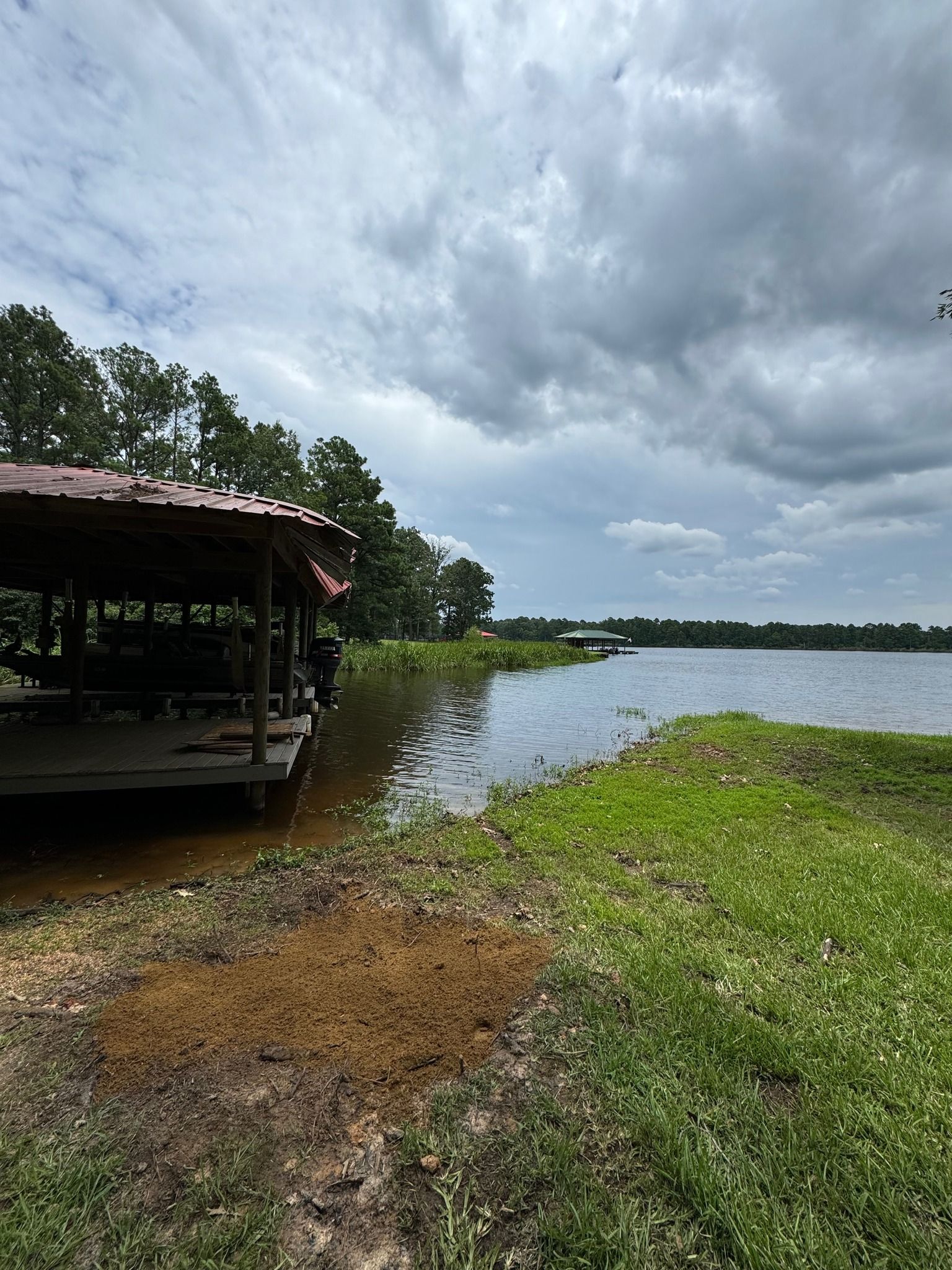 Cloudy lakeside view with a covered dock on the left and grassy shoreline in the foreground.