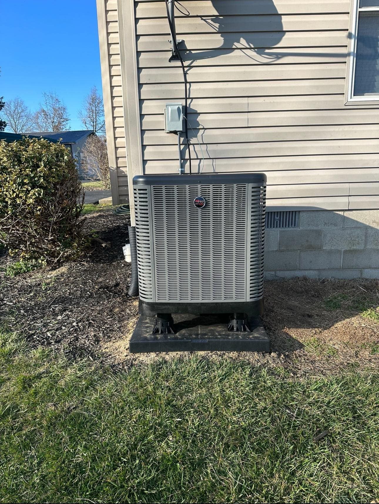 Outdoor HVAC unit beside beige house siding and foundation, with grass and mulch in front.