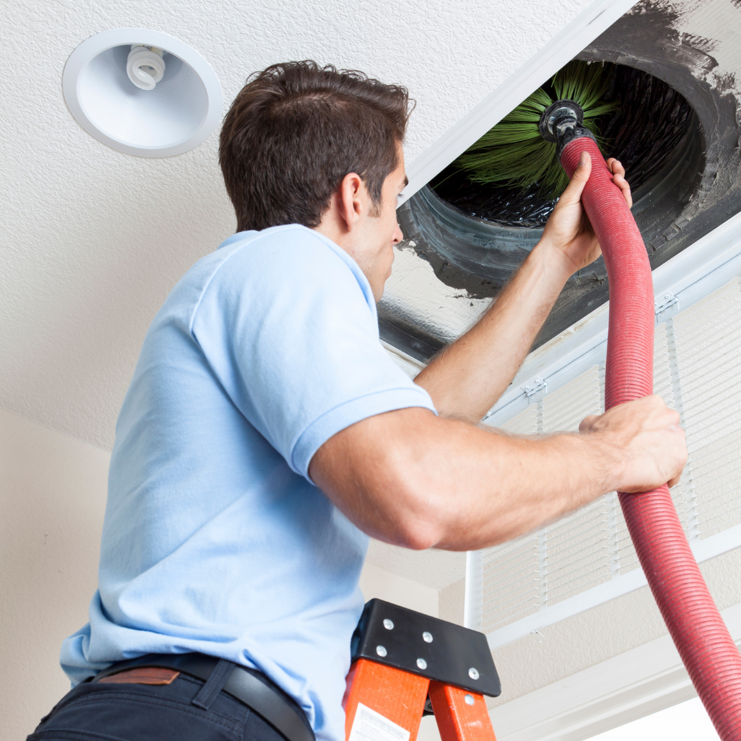 A man on a ladder is cleaning an air vent with a brush