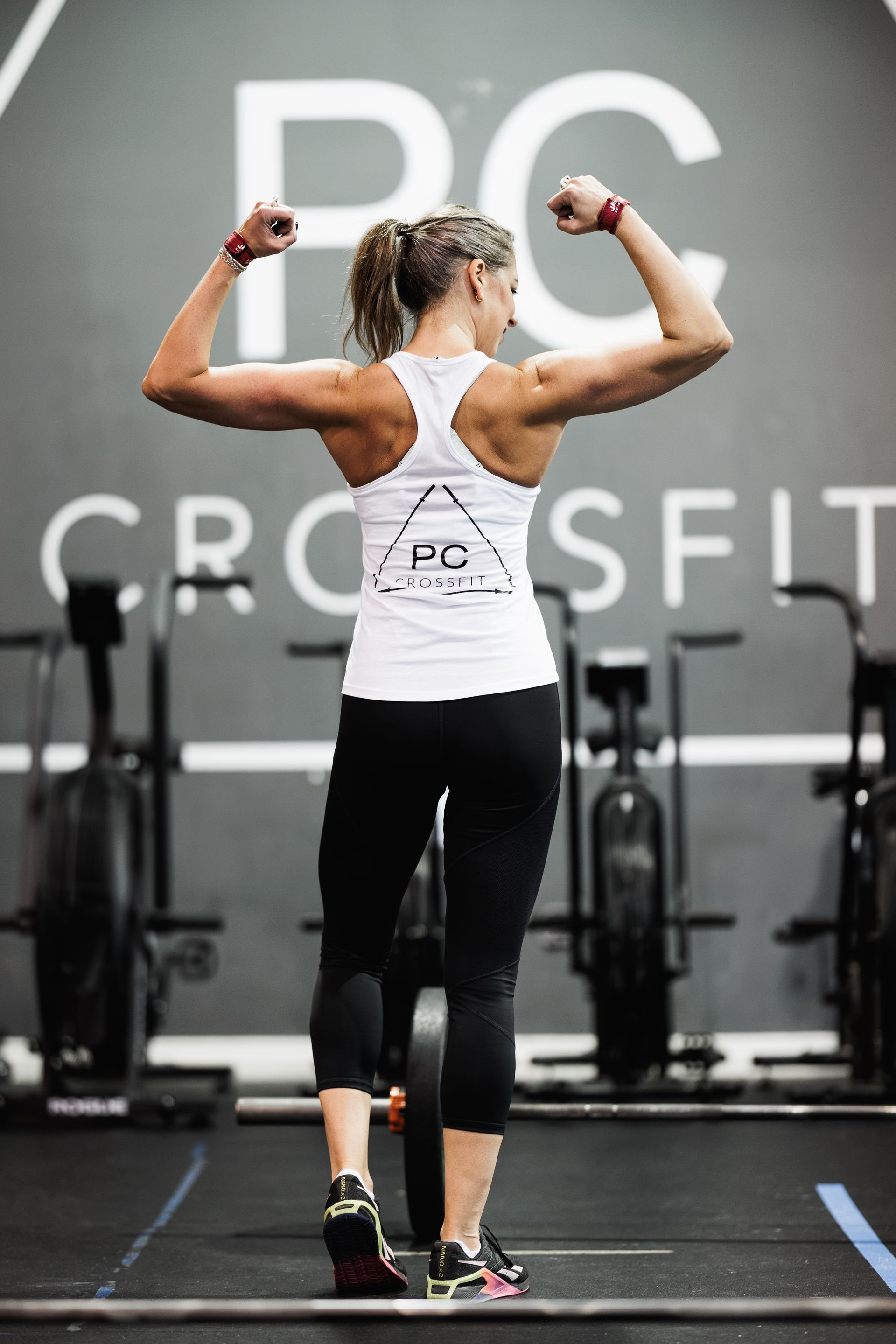 Woman flexes biceps in gym, wearing a white tank top and black leggings, in front of a CrossFit logo.