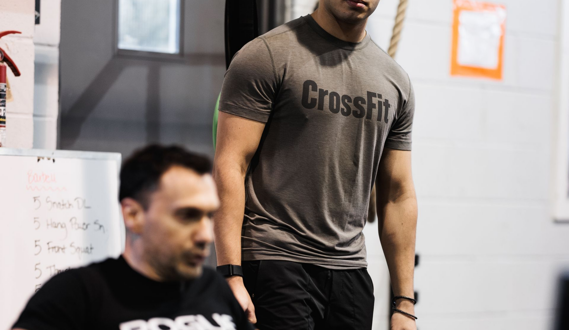 Man watches another row in a CrossFit gym. The rower wears black gloves and a Rogue shirt.
