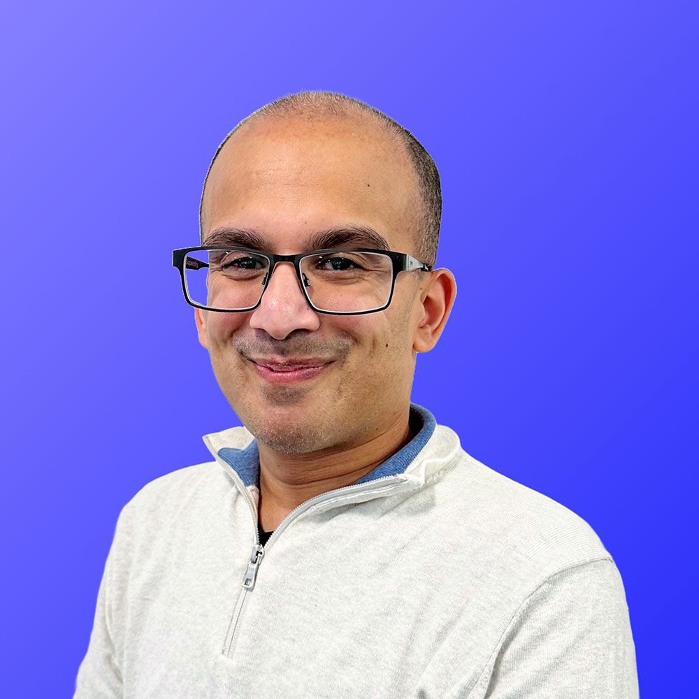 A man wearing glasses and a white sweater is smiling in front of a blue background.