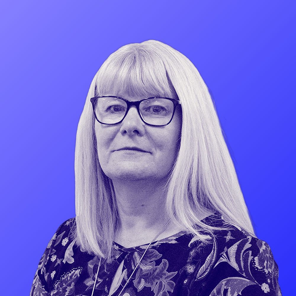 A woman wearing glasses and a floral shirt is standing in front of a blue background.