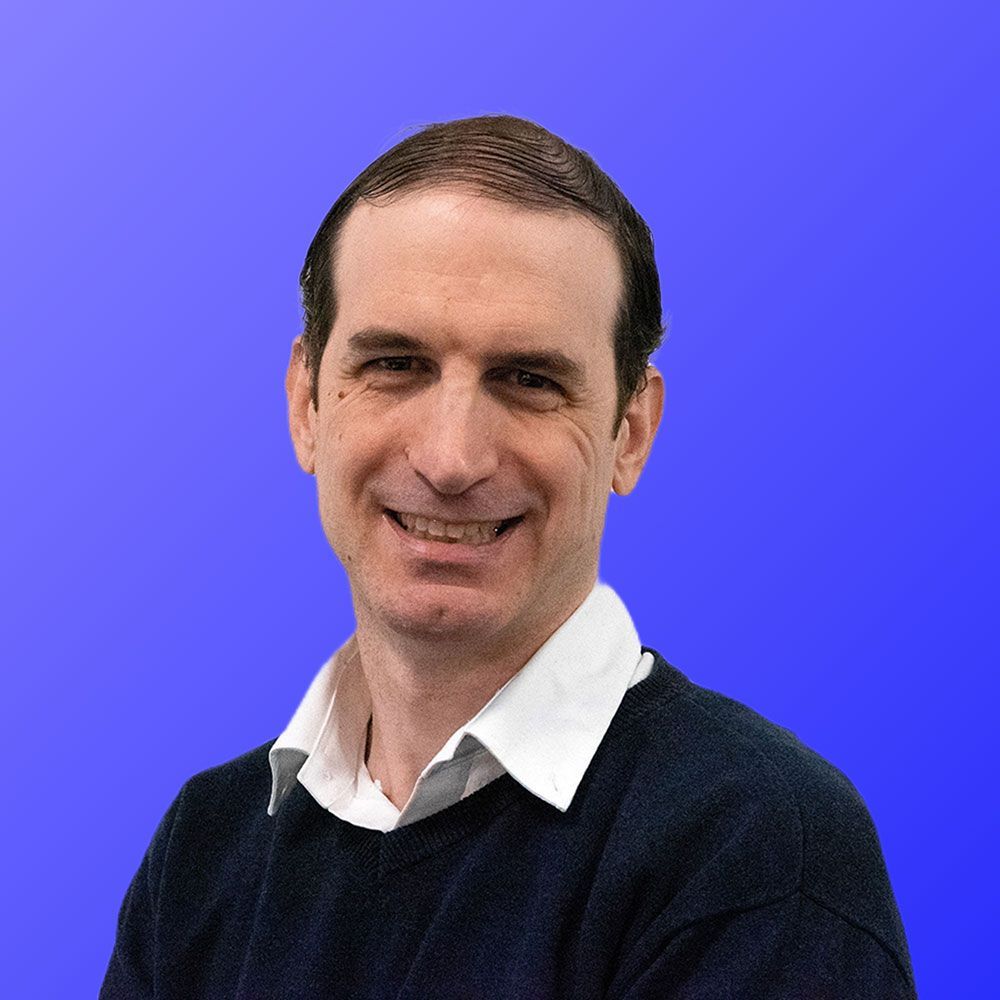 A man in a black sweater and white shirt is smiling in front of a blue background.