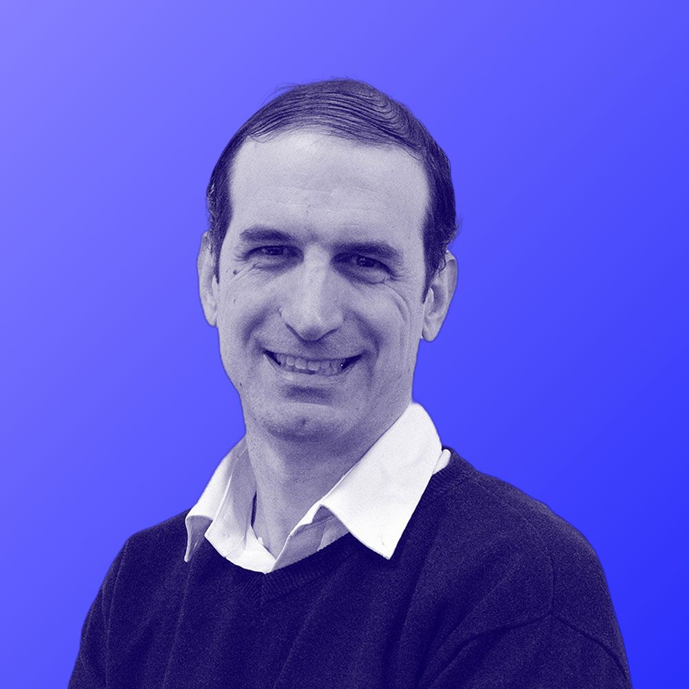 A man in a black sweater and white shirt is smiling in front of a blue background.
