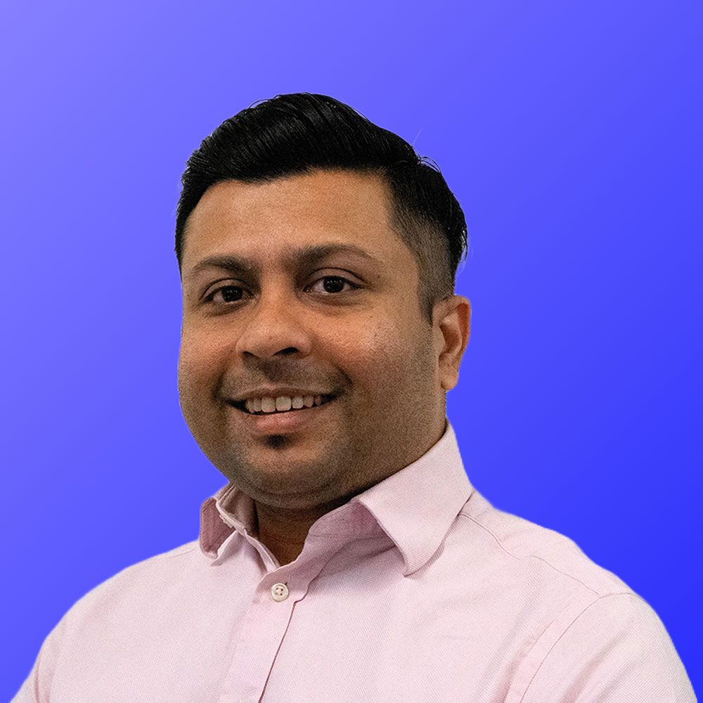 A man in a pink shirt is smiling in front of a blue background.